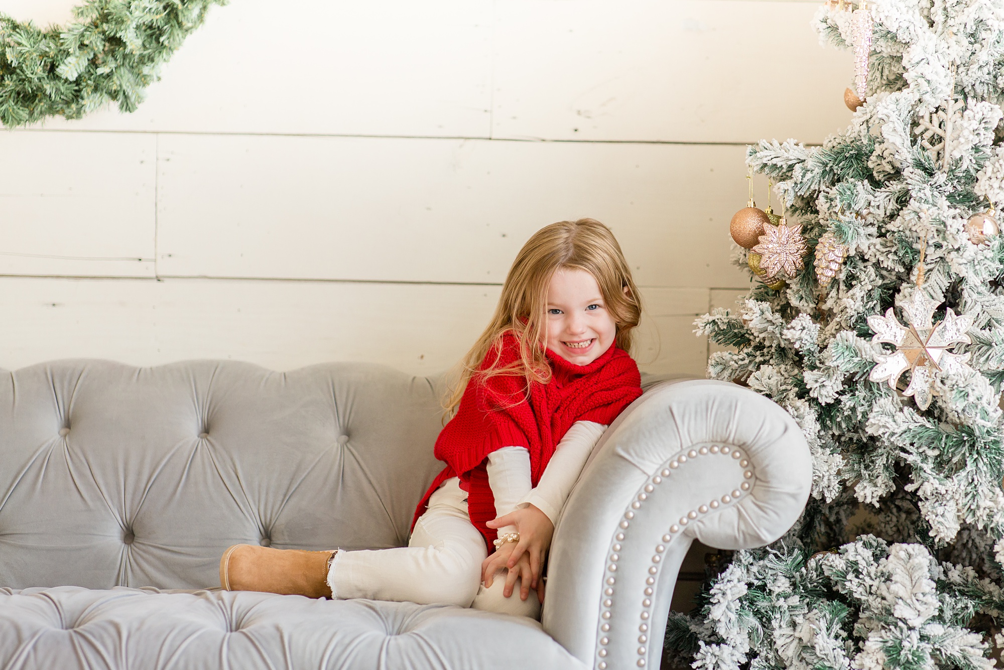 little girl in red sweater sits on grey couch by Christmas tree