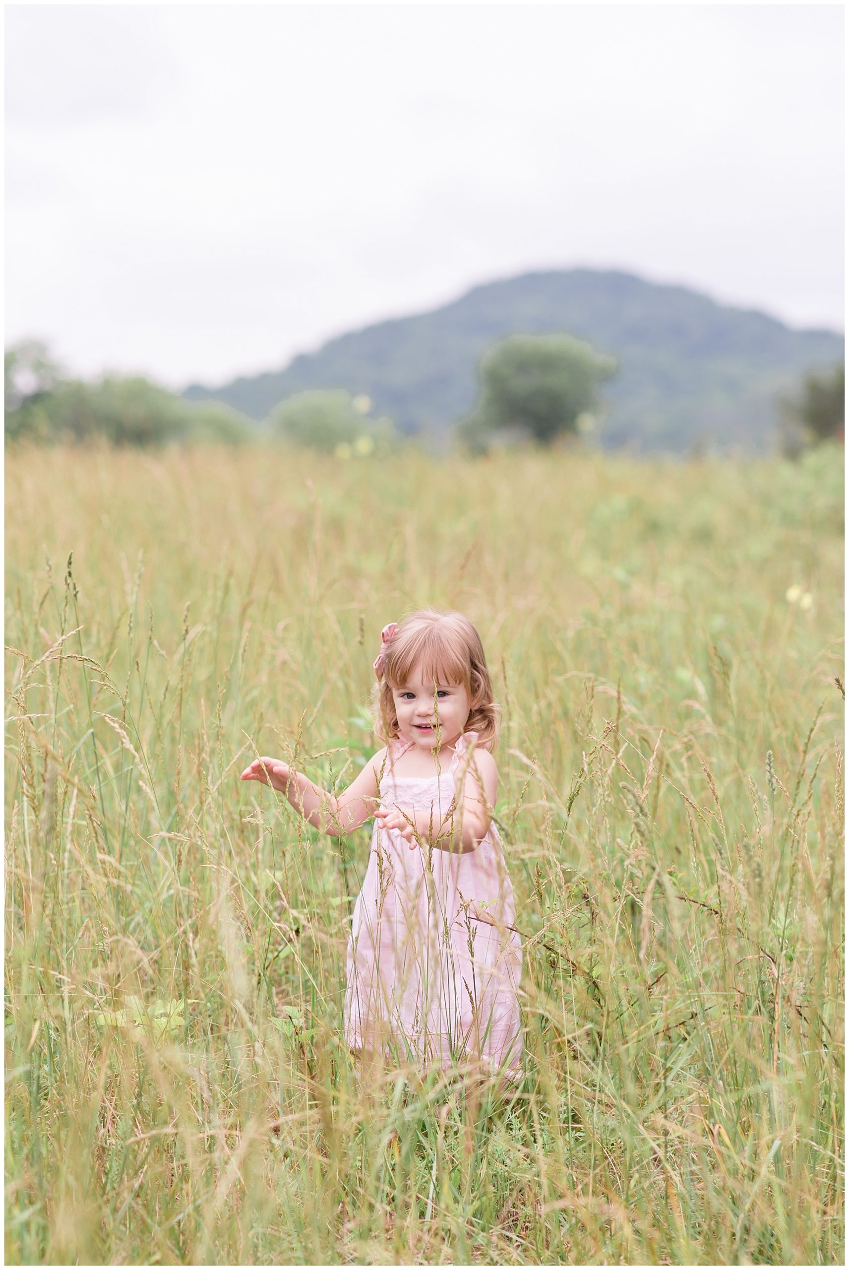 This adorable little girl is seen here playing in a beautiful field. She is wearing a sleeveless light pink dress and a matching pink hair bow.
