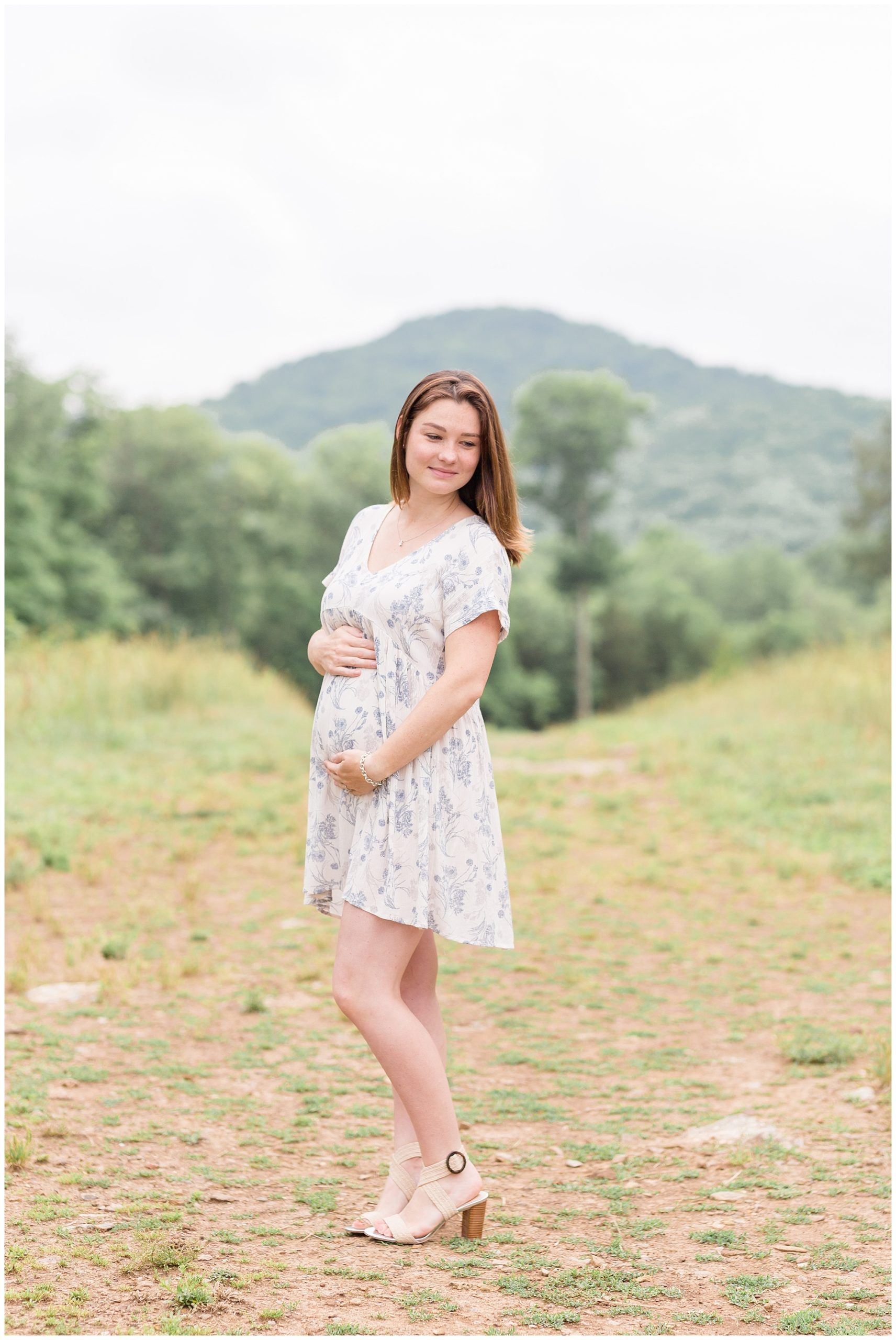 This beautiful mom to be is standing with gorgeous trees and mountians just in the background. She is wearing short sleeve knee length white dress with blue floral print and lace detail at the neckline.