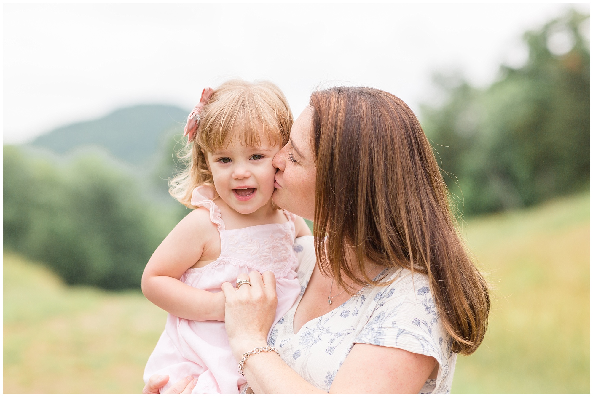 Mom is holding daughter and kissing her cheek. Daughter is wearing a soft pink sleeveless dress with matching hair bow. Mom is wearing short sleeve white dress with blue floral print.