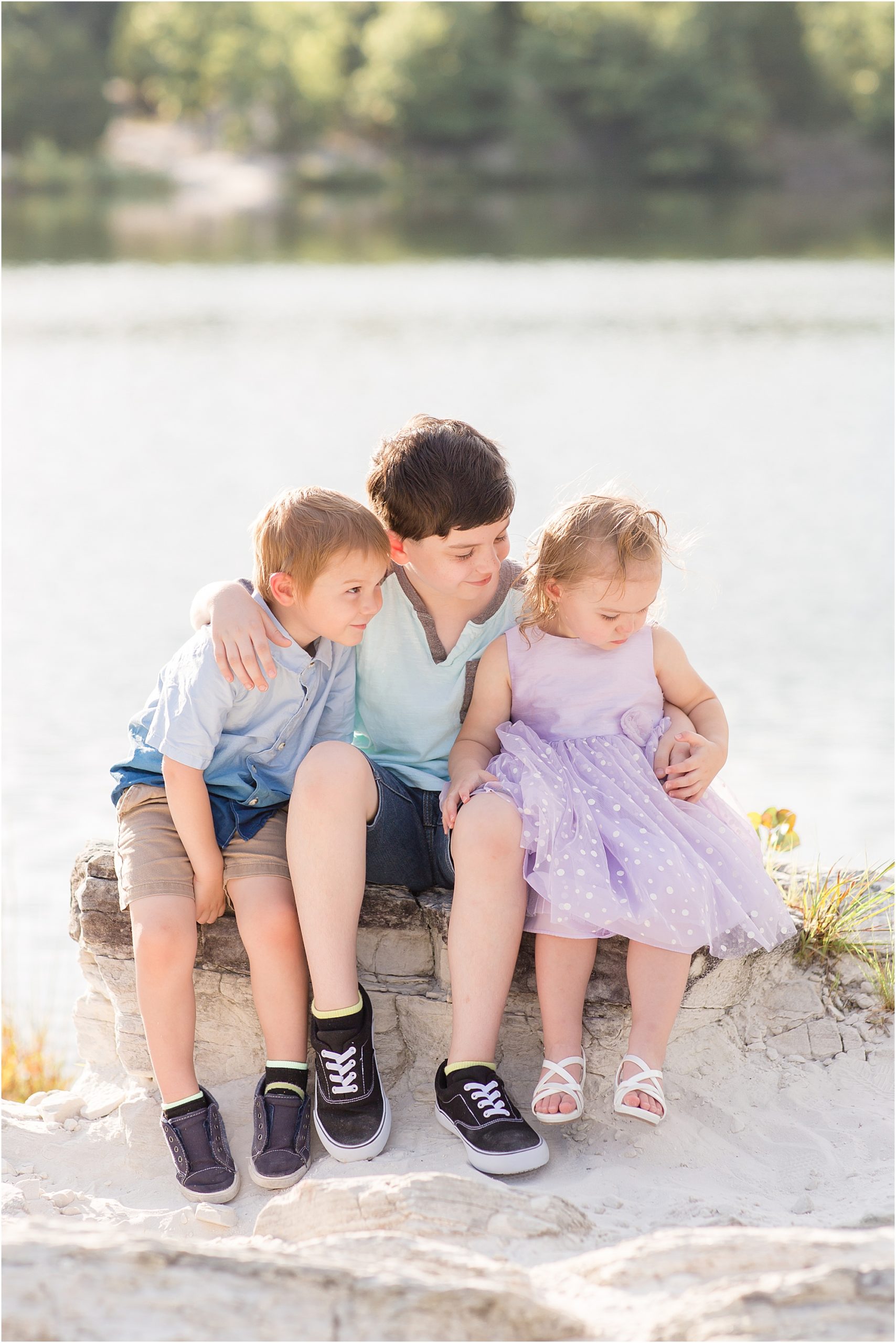 Siblings are seated on a rock just in front of a beautiful lake. 1st brother is wearing a blue and brown jersey style shirt and jeans shorts. 2nd short sleeve button up shirt in shades of blue and khaki shorts. Little sister is wearing a purple dress and white sandals. 