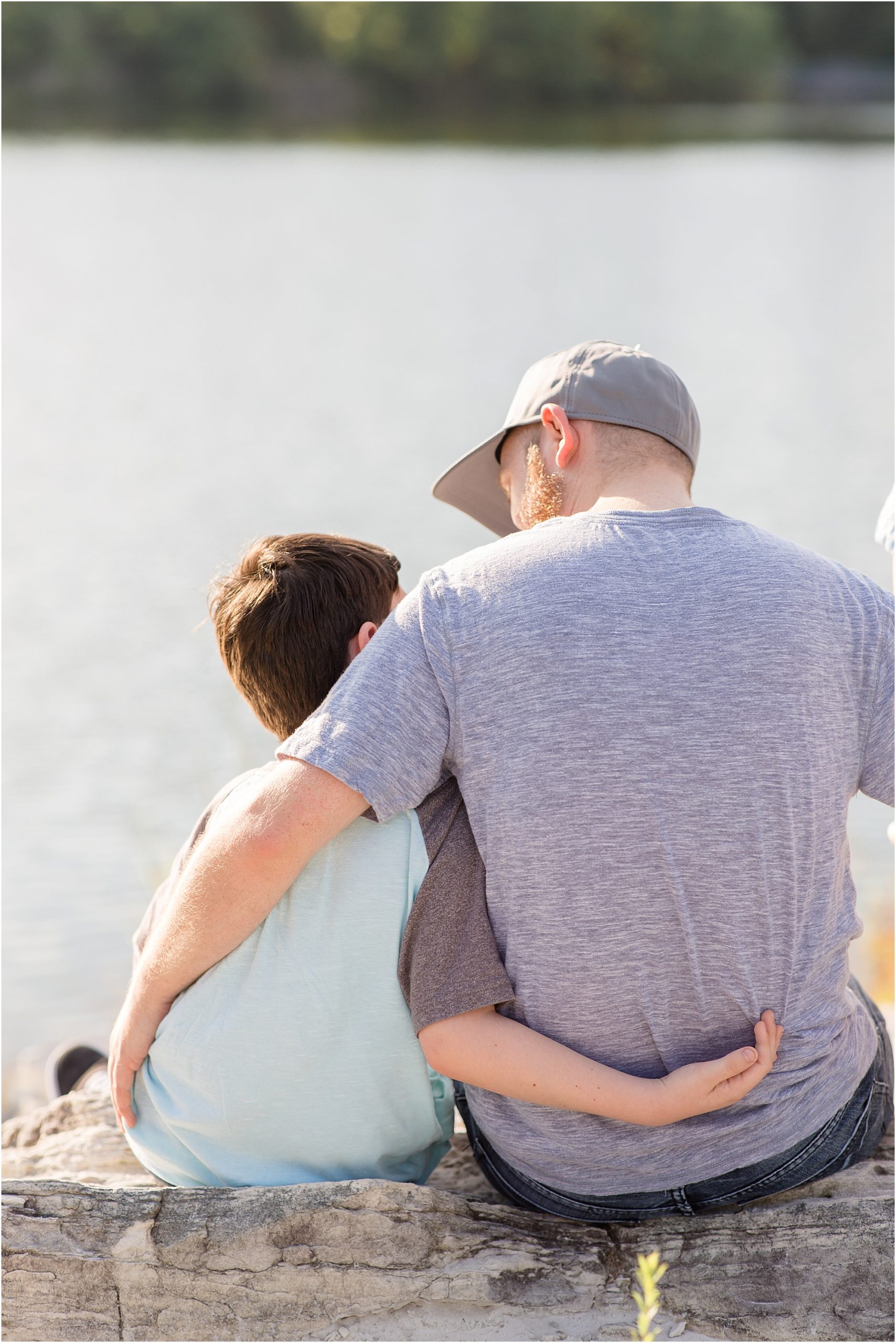 Father and son are seated on rock just in front of a beautiful lake. Dad is wearing a blue shirt with a blue hat and blue jeans. Son is wearing a blue and brown jersey style shirt and jeans shorts. 