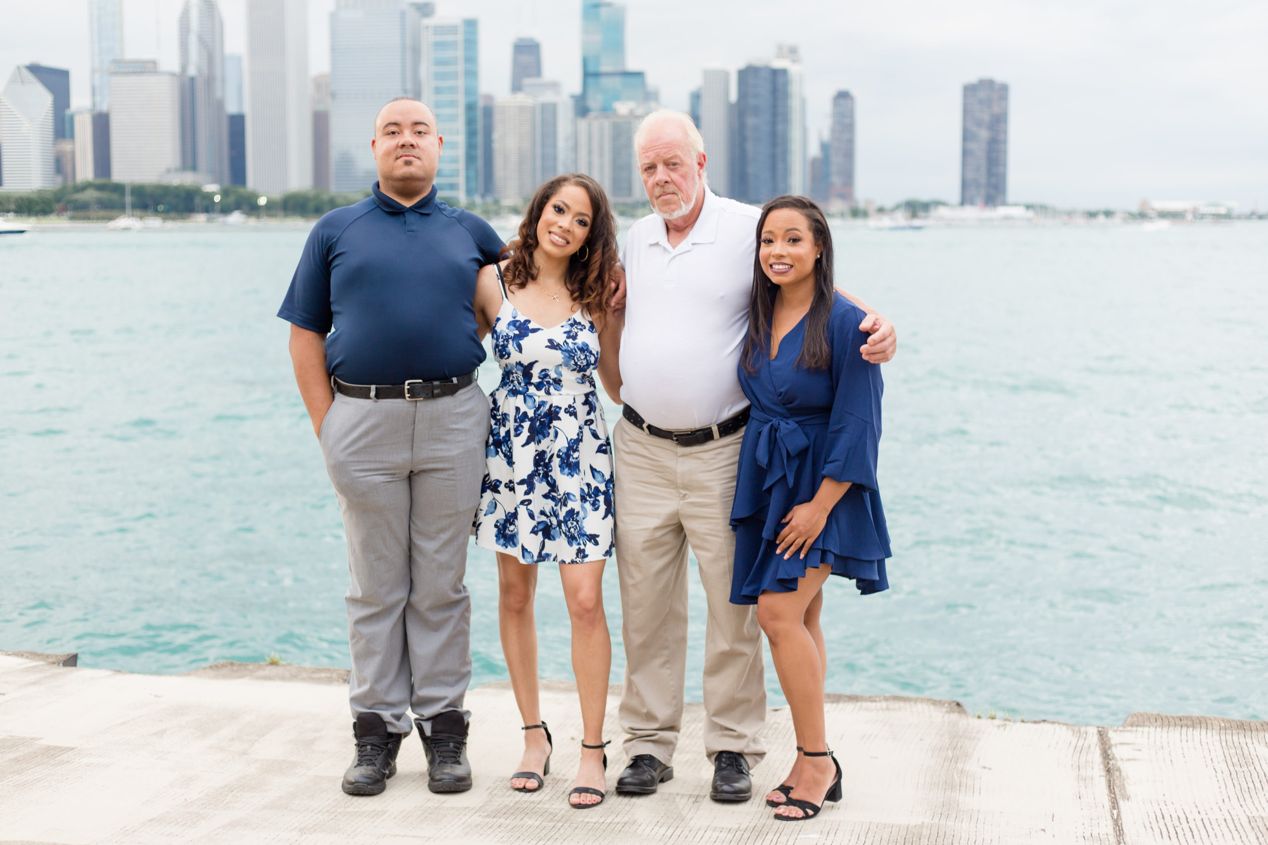 Family of 4 standing with Chicago skyline in the back. Son is wearing a navy blue polo and grey pants. 1st daughter is wearing a sleeveless blue and white floral print dress. Dad is wearing a white polo and khaki pants. 2nd daughter is wearing a long sleeve navy blue dress. 