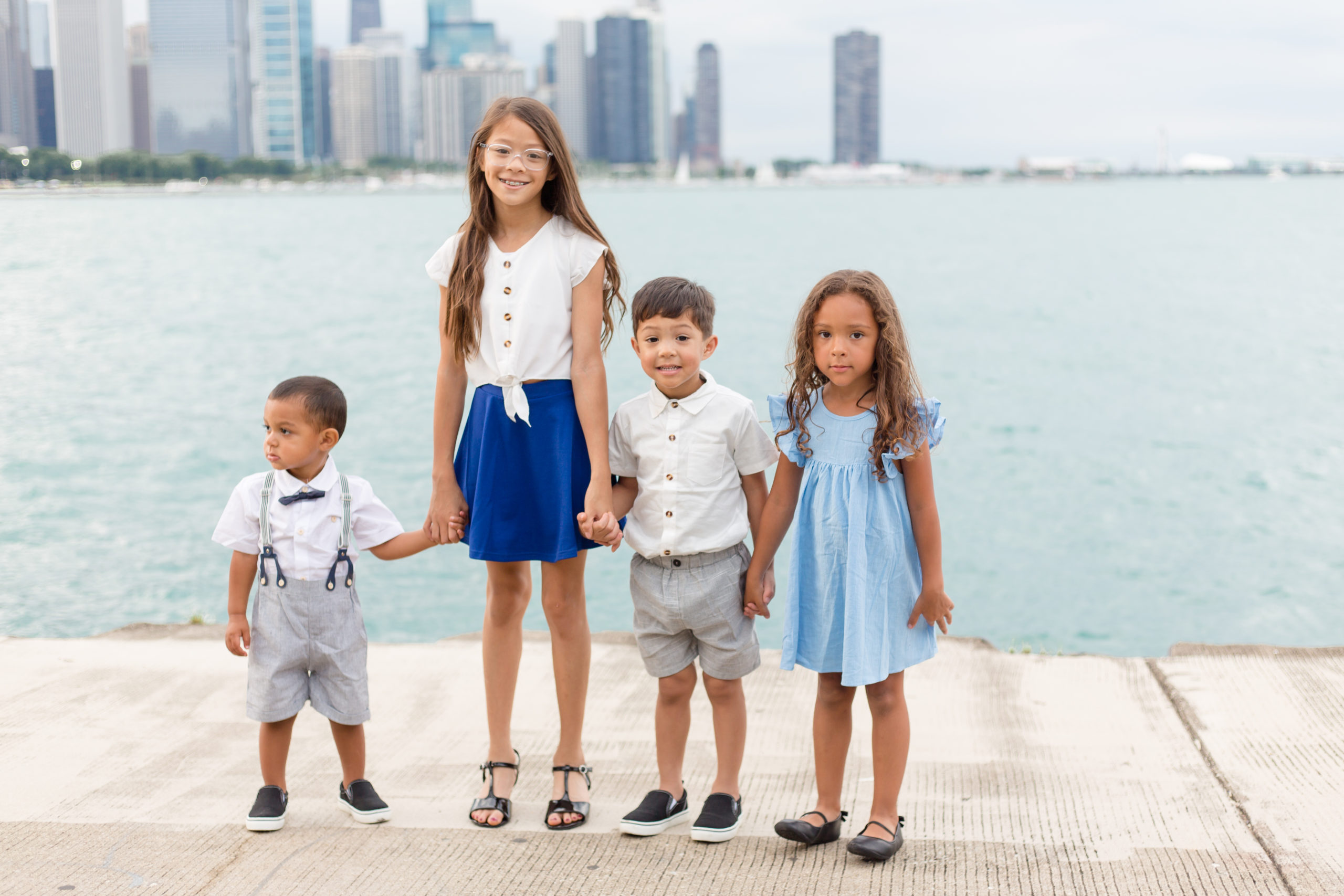 These 4 cousins are standing holding hands in front of a beautiful lake. 1st cousin is wearing a white shirt, bow tie, suspenders and grey shorts. 2nd cousin is wearing a white shirt that ties at the wait and a royal blue skirt. 3rd cousin is wearing a white button up shirt and grey shorts. 4th cousin is wearing a light blue flutter sleeve dress. Check out more about this gorgeous extended family session in the blog.