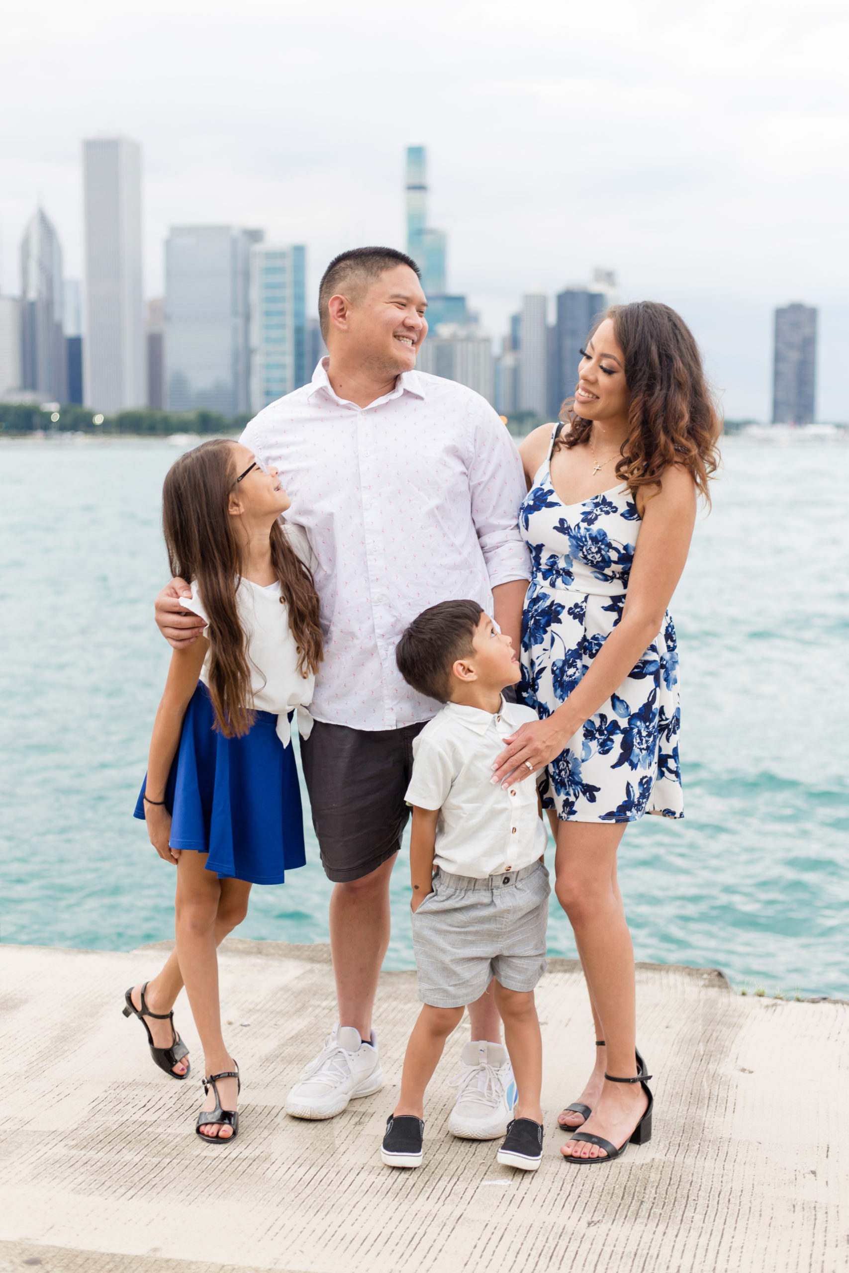 Beautiful family of 4 standing with the Chicago skyline in the back. Little girl is dressed in a white top that ties at the waist and a vibrant blue skirt. Dad is wearing a white button up shirt and grey shorts. Mom is in a sleeveless dress with blue and white floral design. Son is dressed in a white button up shirt and grey shorts.