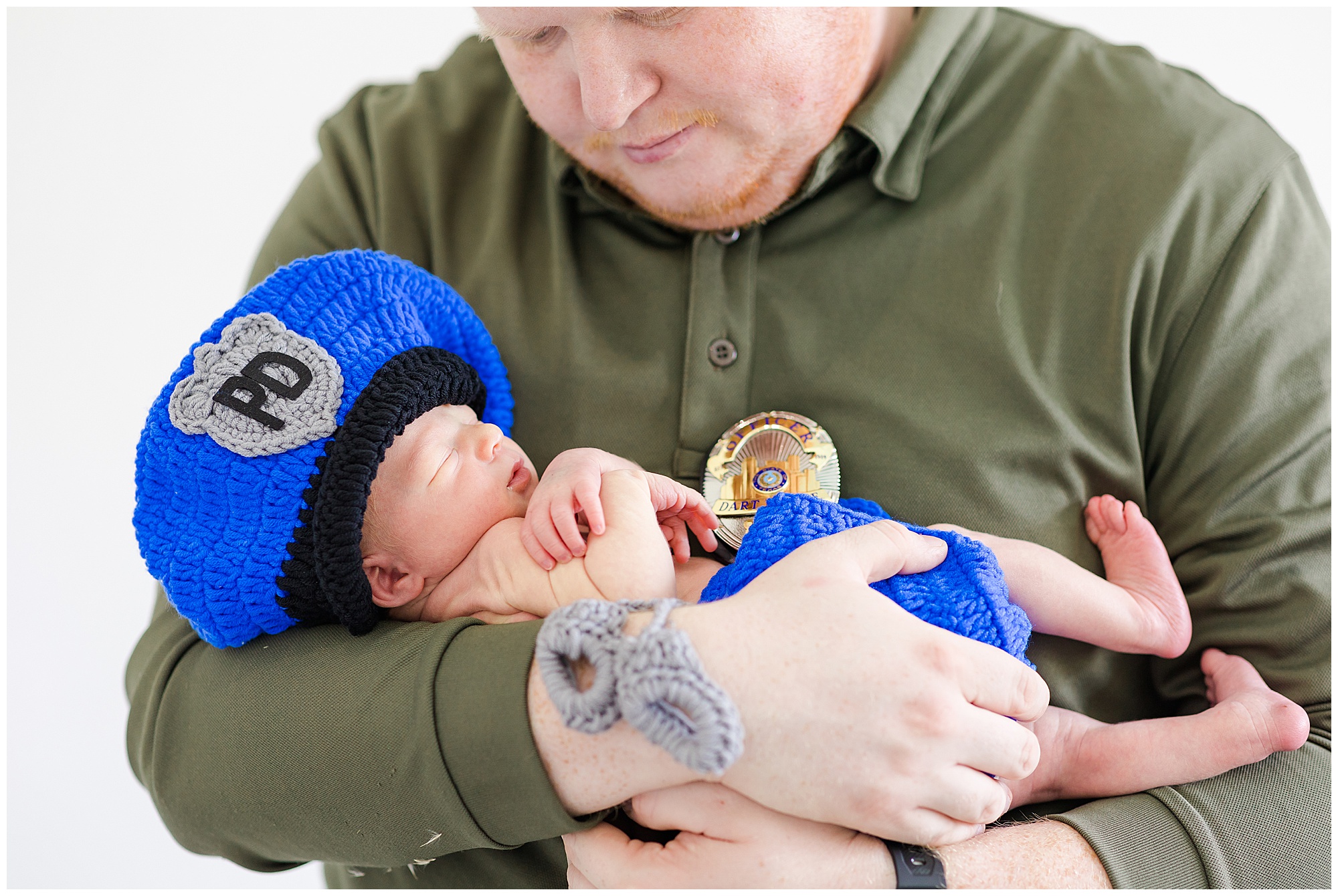 Dad in dark green long sleeve shirt holds newborn infant in knit baby police outfit during lifestyle newborn session in McKinney, TX. 