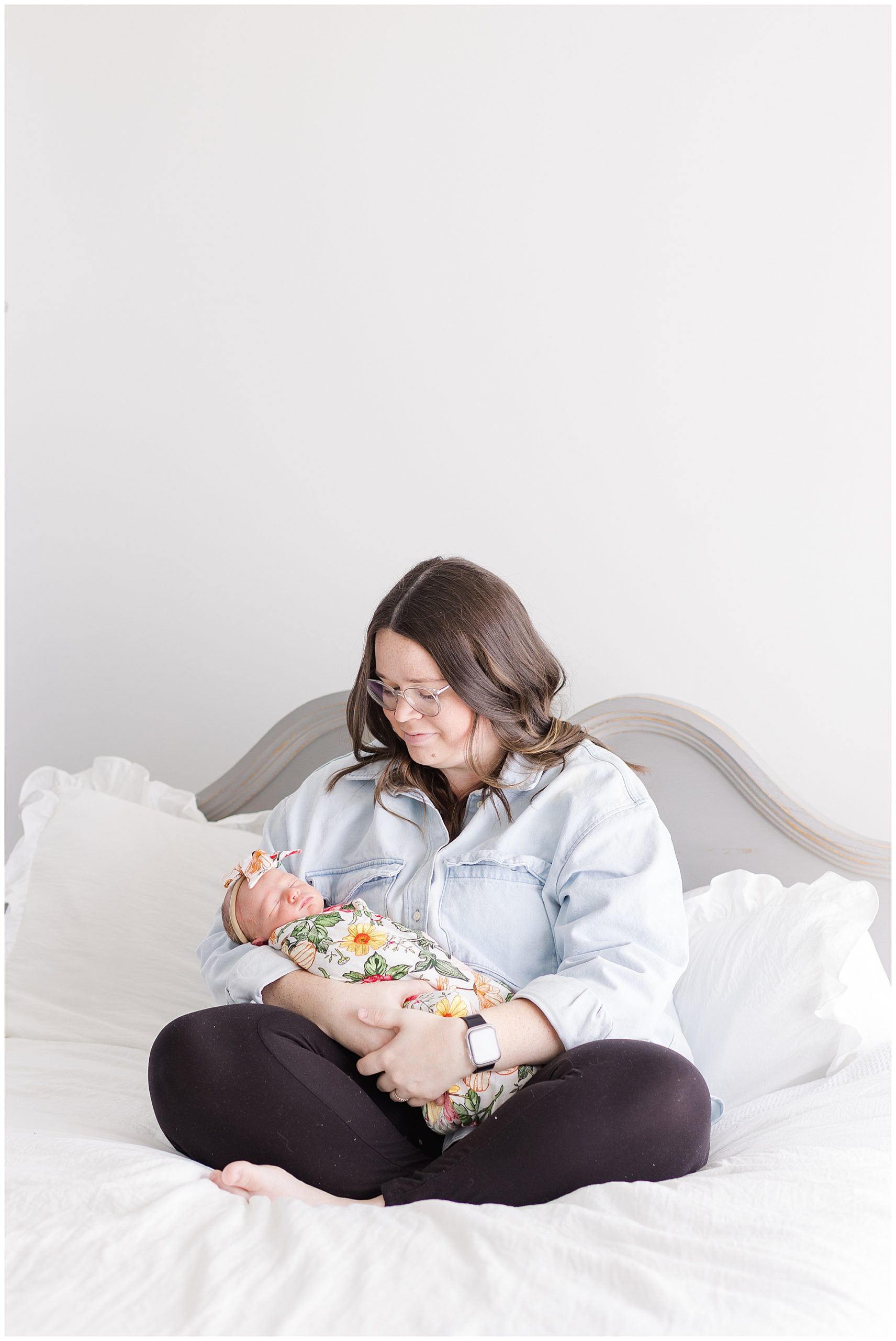 Mom in light blue button down and black pants sits on bed with white linens while holding newborn daughter in colorful floral swaddle and bow during lifestyle newborn session. 