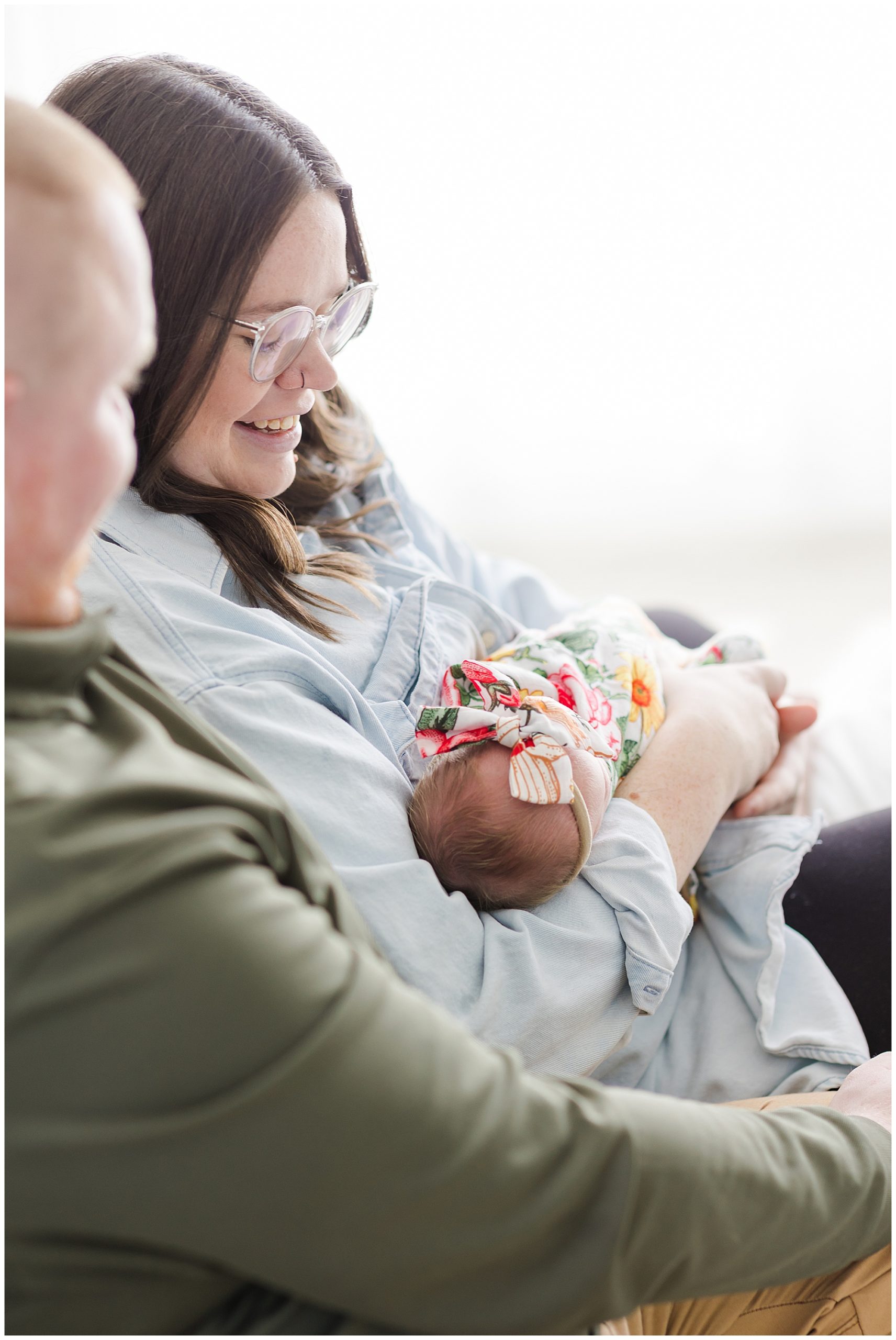 Mom in light blue button down and dad in green shirt sit together while holding newborn daughter in colorful swaddle during lifestyle newborn session in McKinney, TX. 