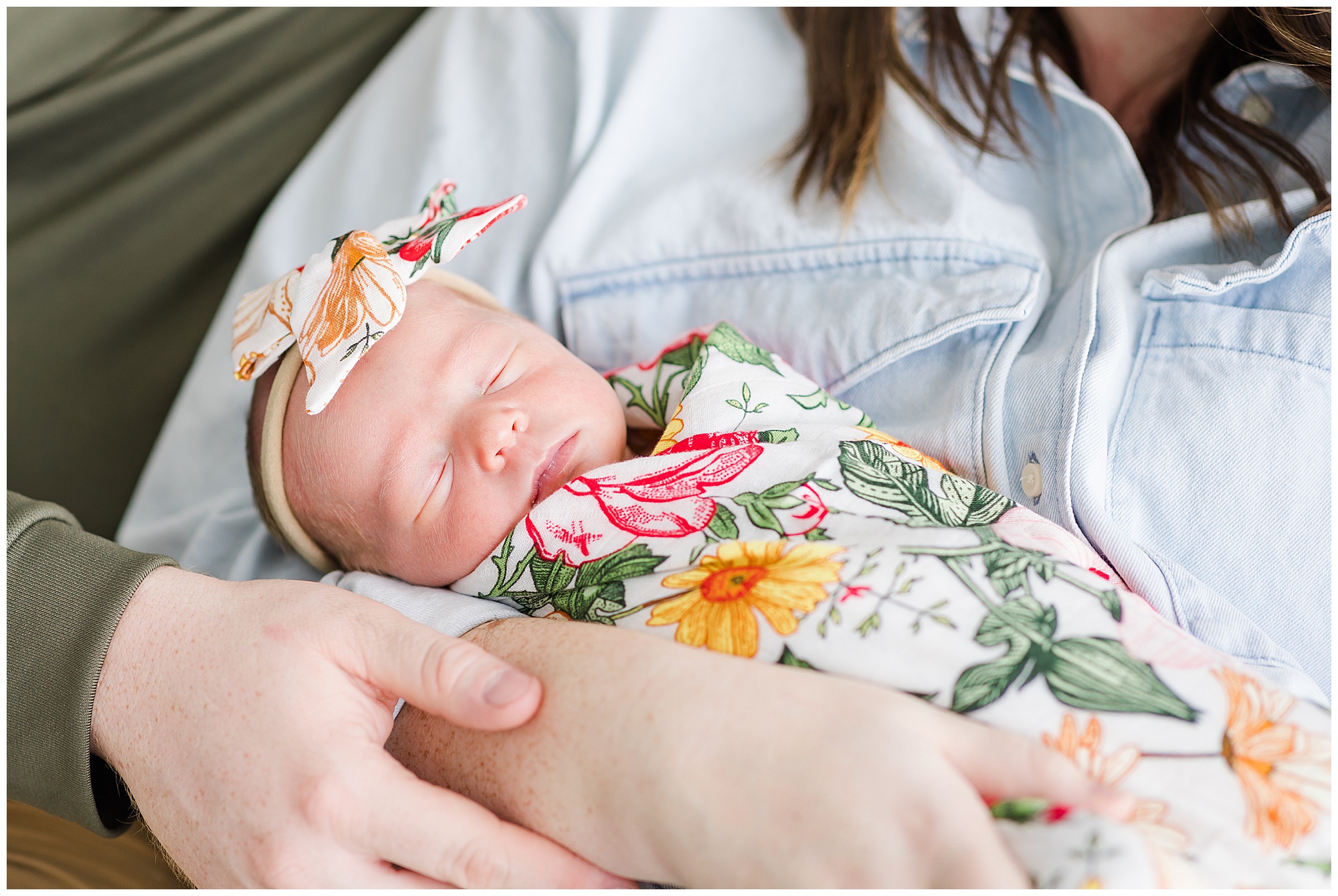 Newborn baby sleeps in mom's arms while wearing colorful floral swaddle and bow during Behind the Lens lifestyle newborn session. 
