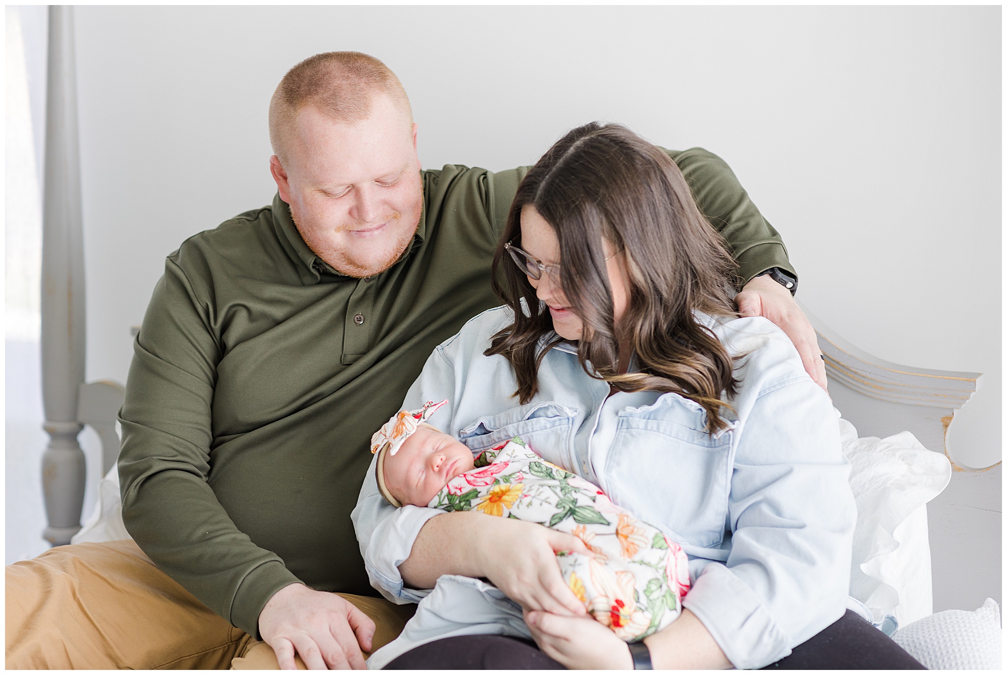 Dad in dark green shirt and mom in light blue shirt hold sweet newborn daughter in colorful floral swaddle during Behind the Lens session. 