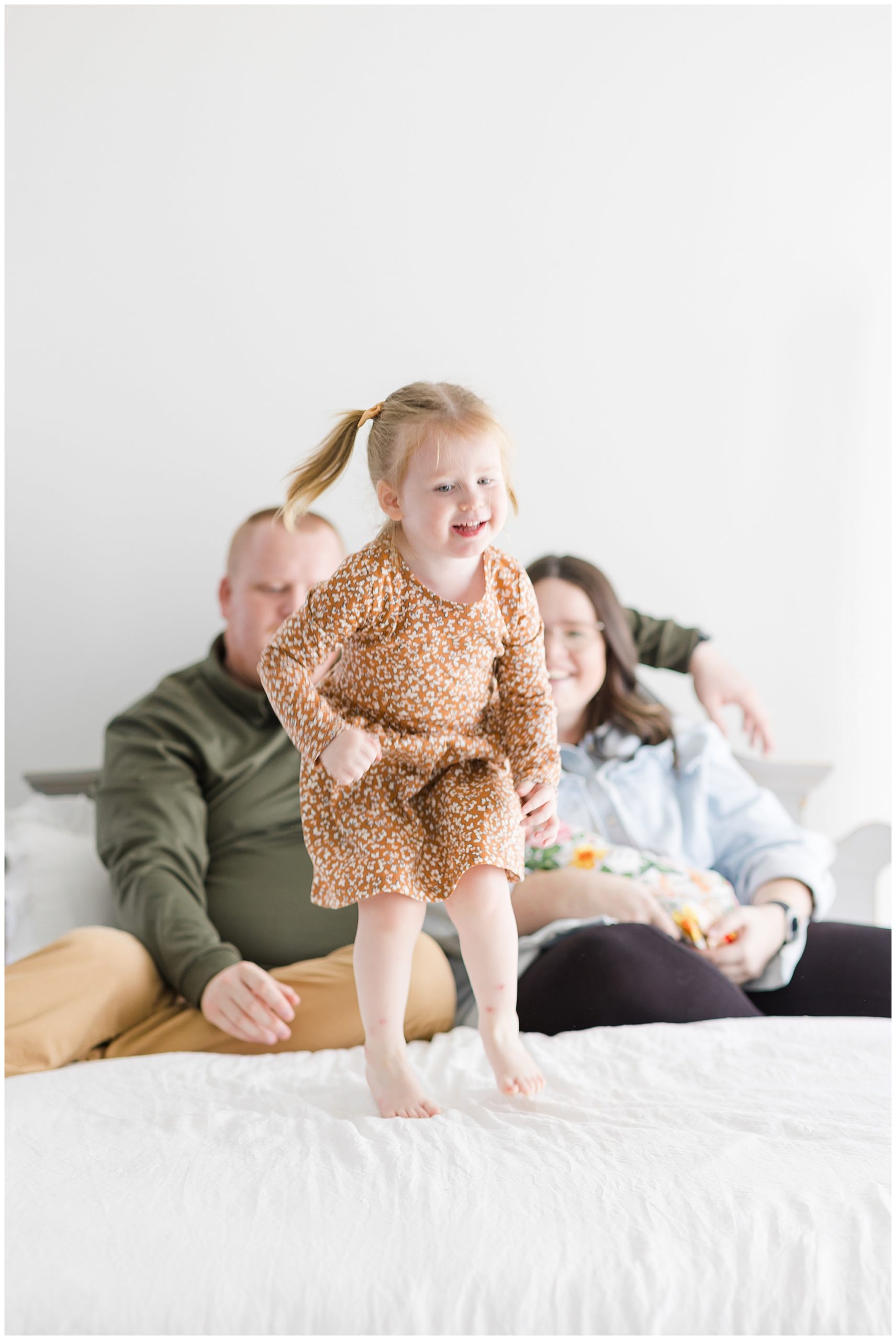 Little girl in orange floral dress jumps on bed with white linens with mom and dad and newborn sister behind her during lifestyle newborn family session. 