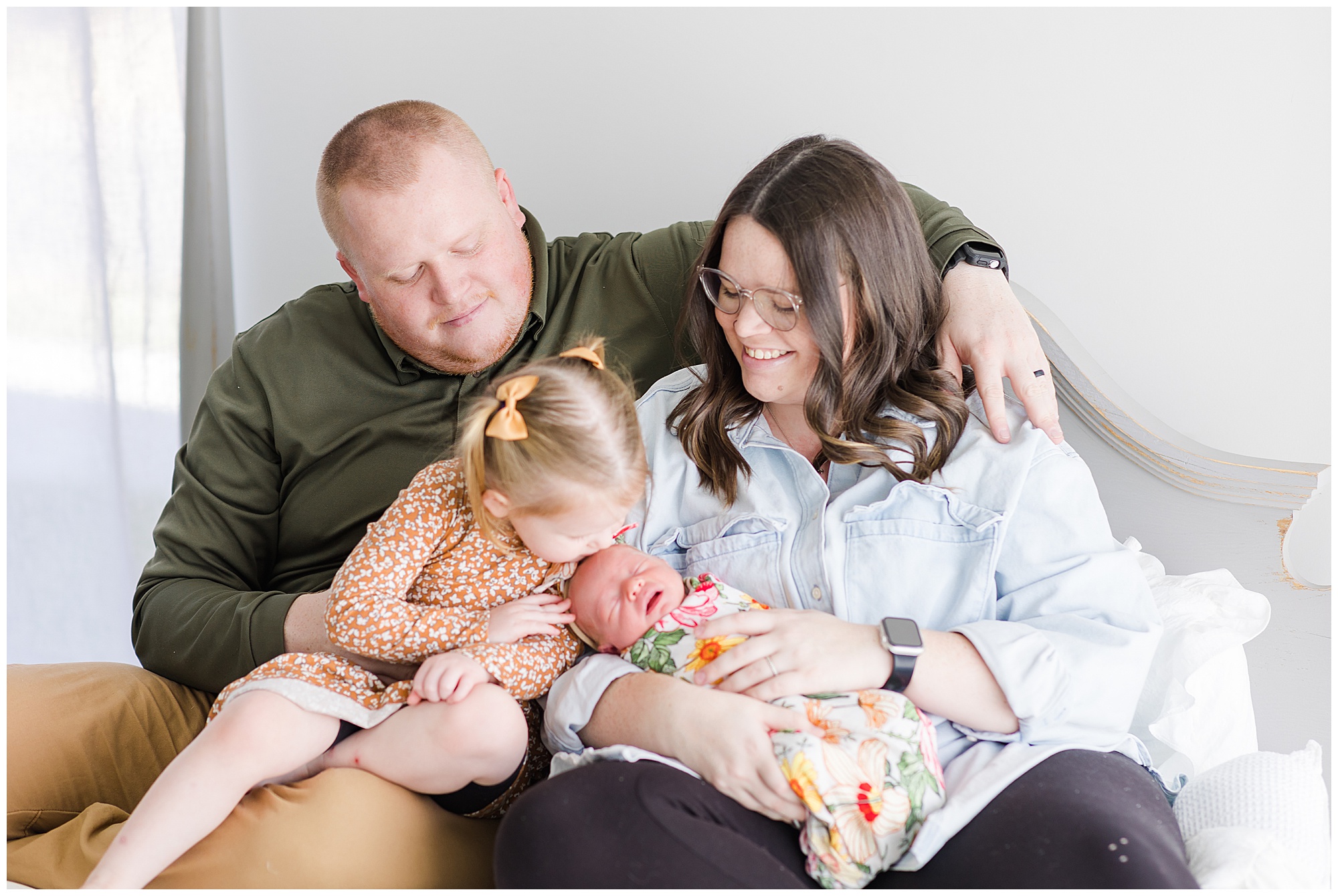 Dad in green shirt and mom in light blue button down sit on white bed while toddler daughter in orange dress kisses baby sister in colorful swaddle during newborn family session. 