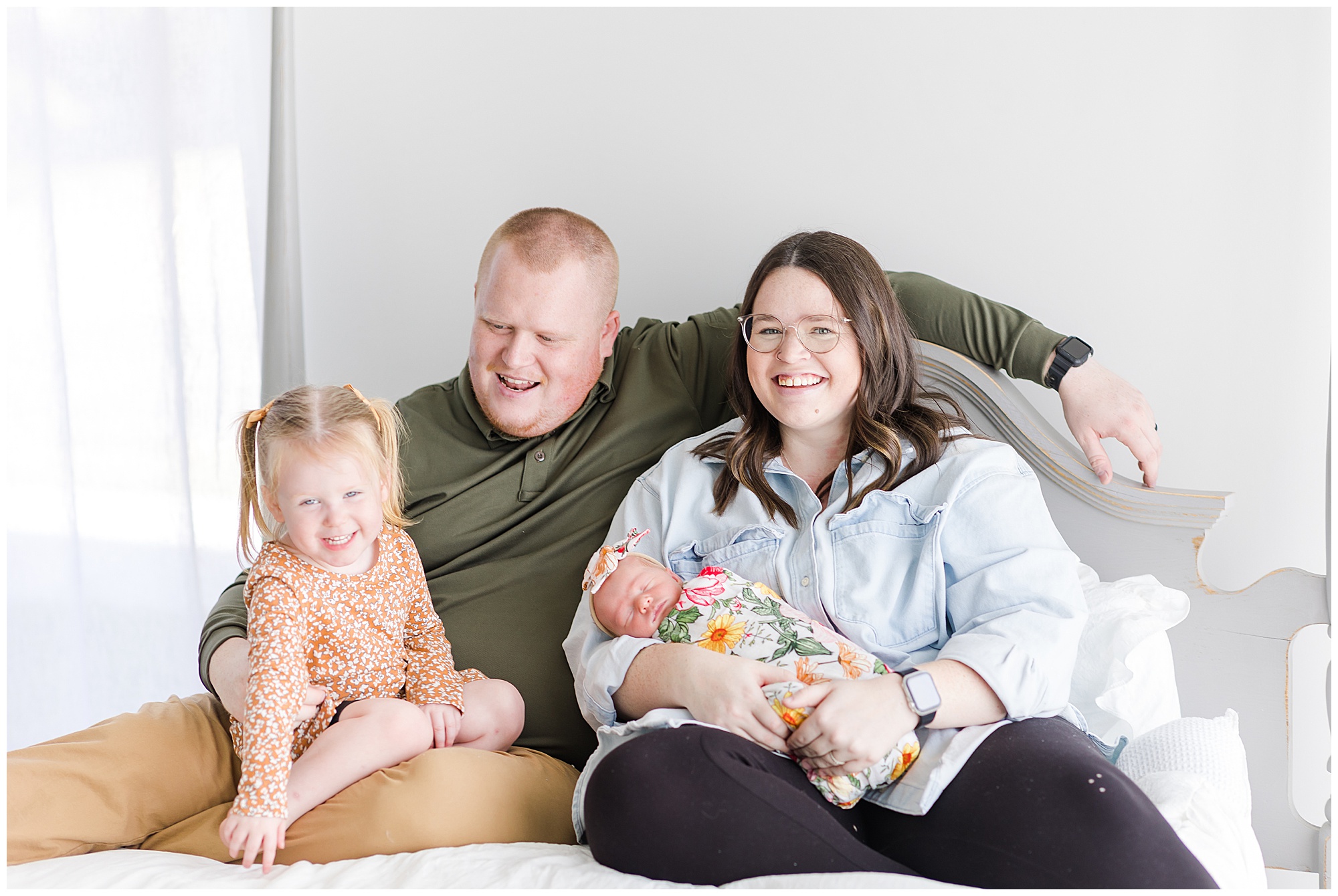 Mom and dad sit on bed with white sheets while holding newborn baby in colorful floral swaddle and toddler in orange dress during family photo session. 