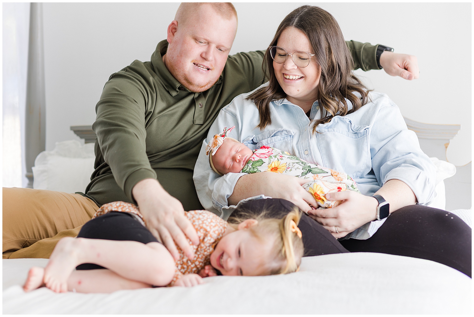 Dad in dark green shirt tickles toddler daughter while mom in light blue shirt holds newborn infant while they sit on a bed with white linens during newborn session. 