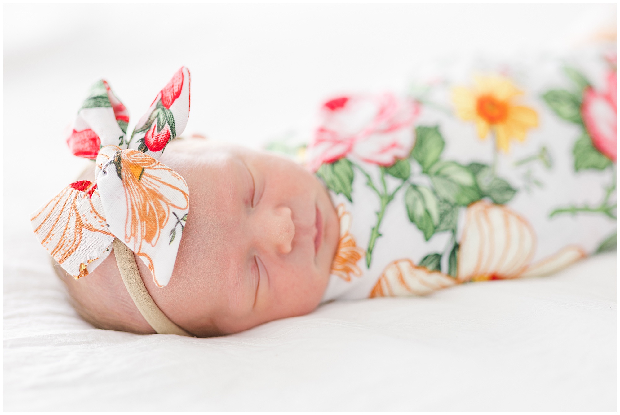 Newborn infant in colorful floral swaddle and bow sleeps on white linens during lifestyle newborn session in McKinney, TX. 