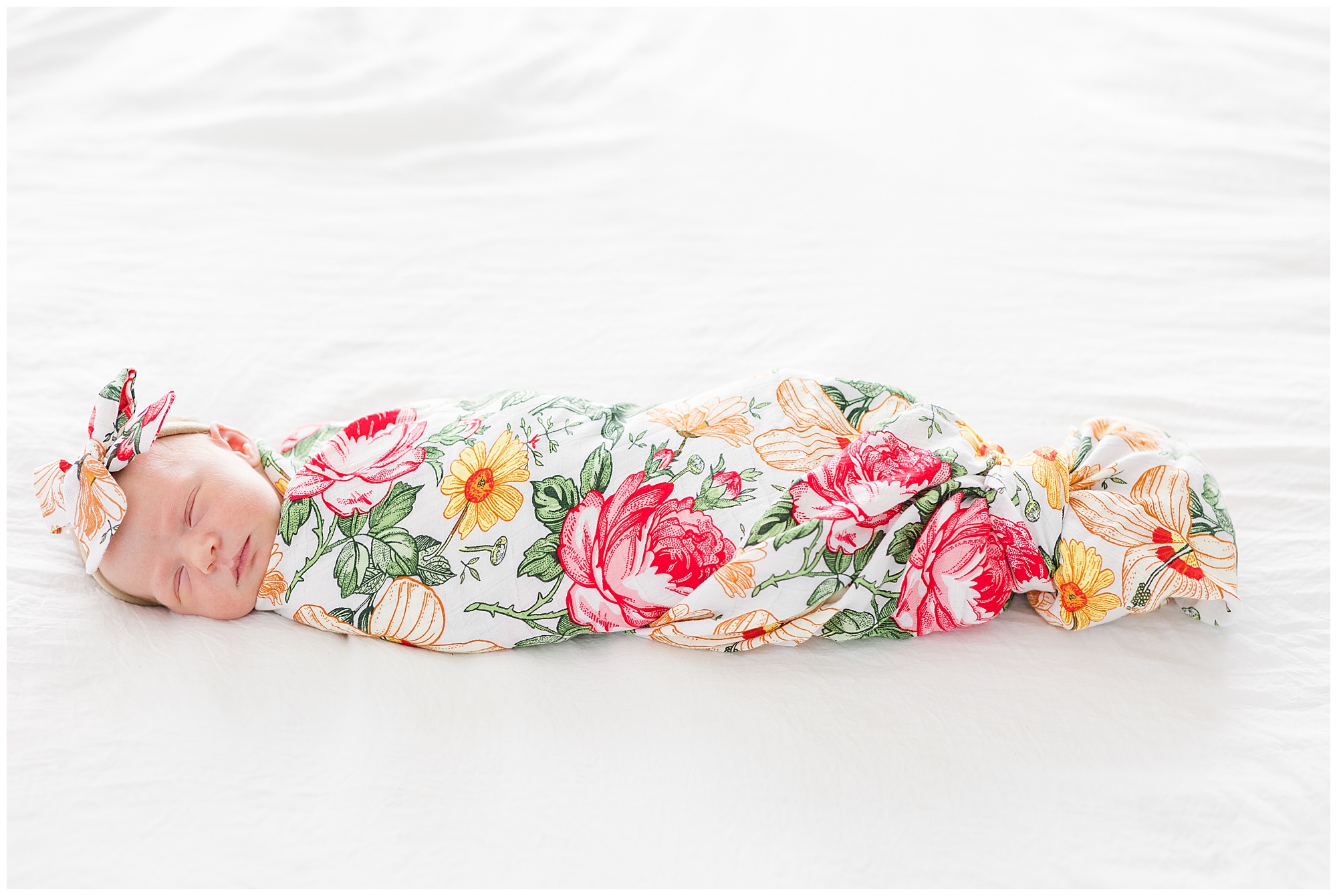 Newborn infant in colorful floral swaddle and bow sleeps on white sheets during lifestyle newborn family session. 