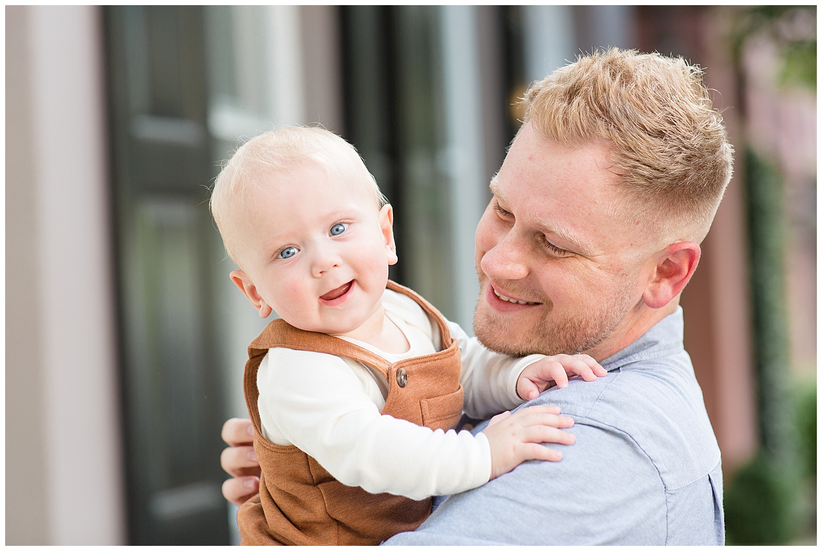 Blonde hair and blue eyed baby boy is being held by his Dad and looking over his shoulder and smiling at the camera of Rebecca Rice Photography during their family session in Charleston, SC.