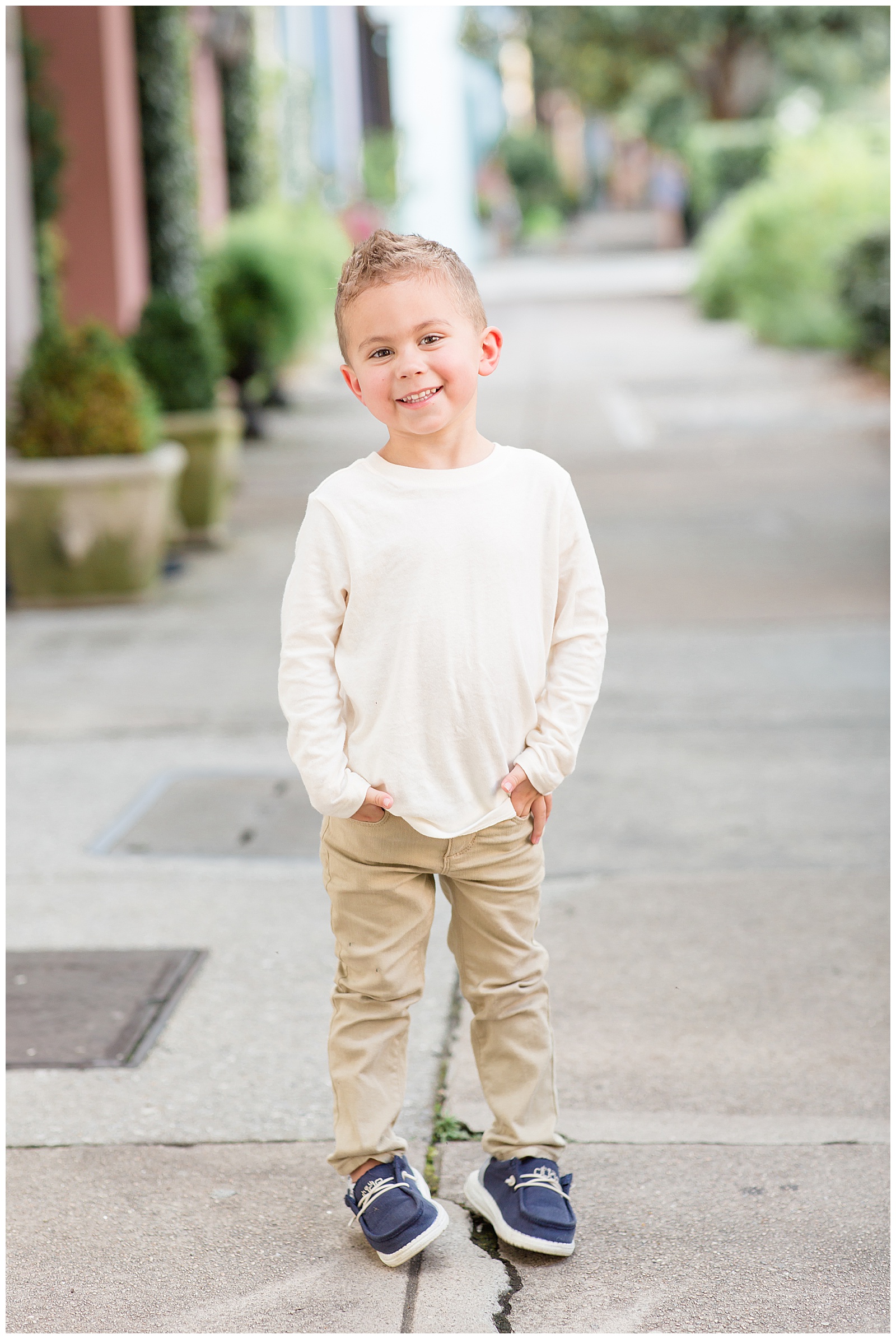 Little boy model stands on the street of Rainbow Row as he wears a cream long sleeve shirt, khakis, and blue shoes.