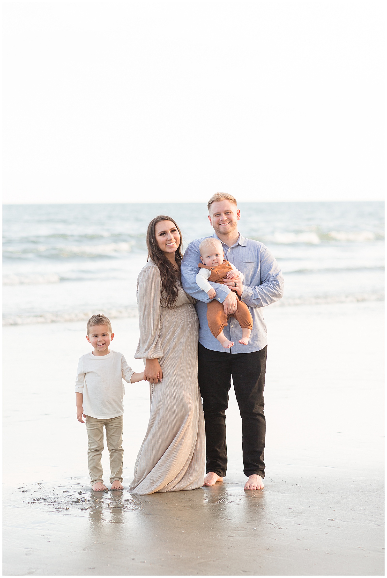Mom and Dad stand together with two sons on the beaches of Charleston as family photographer, Rebecca Rice, captures their smiles. Family wears coordinating outfits of neutral colors.