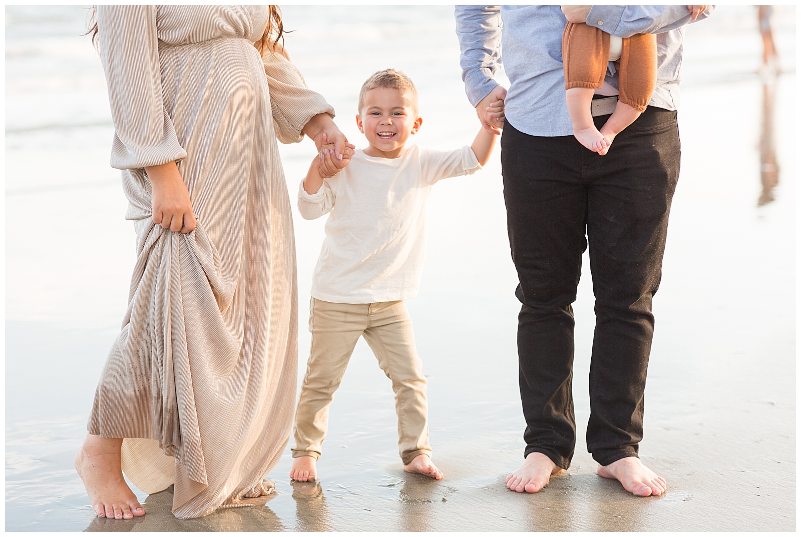 Mom and Dad hold hands of son as they walk together on the Isle of Palms Beach in Charleston, SC as he smiles at the camera and shares his pure joy.