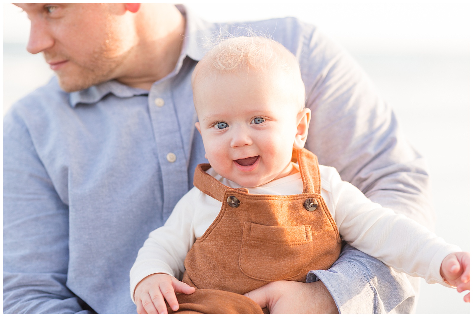Blue-eyed, blond hair little boy smiles at camera in his brown overalls as his Dad holds him in his arms.