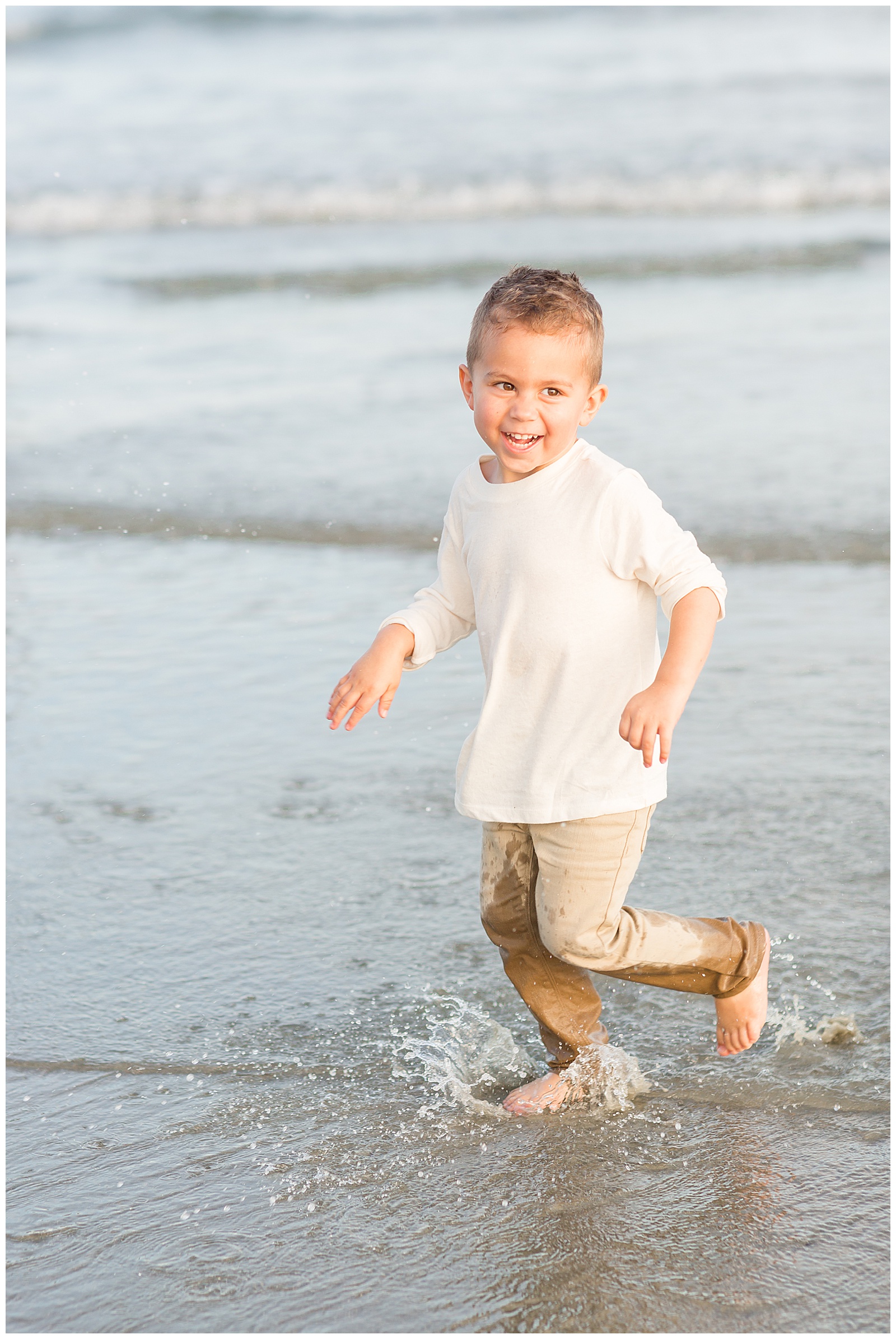 Little boy shares joyful moment as he runs in the waters at the beach in Charleston, SC wearing khaki pants and a cream long sleeve shirt during his family photography session with Rebecca Rice Photography.