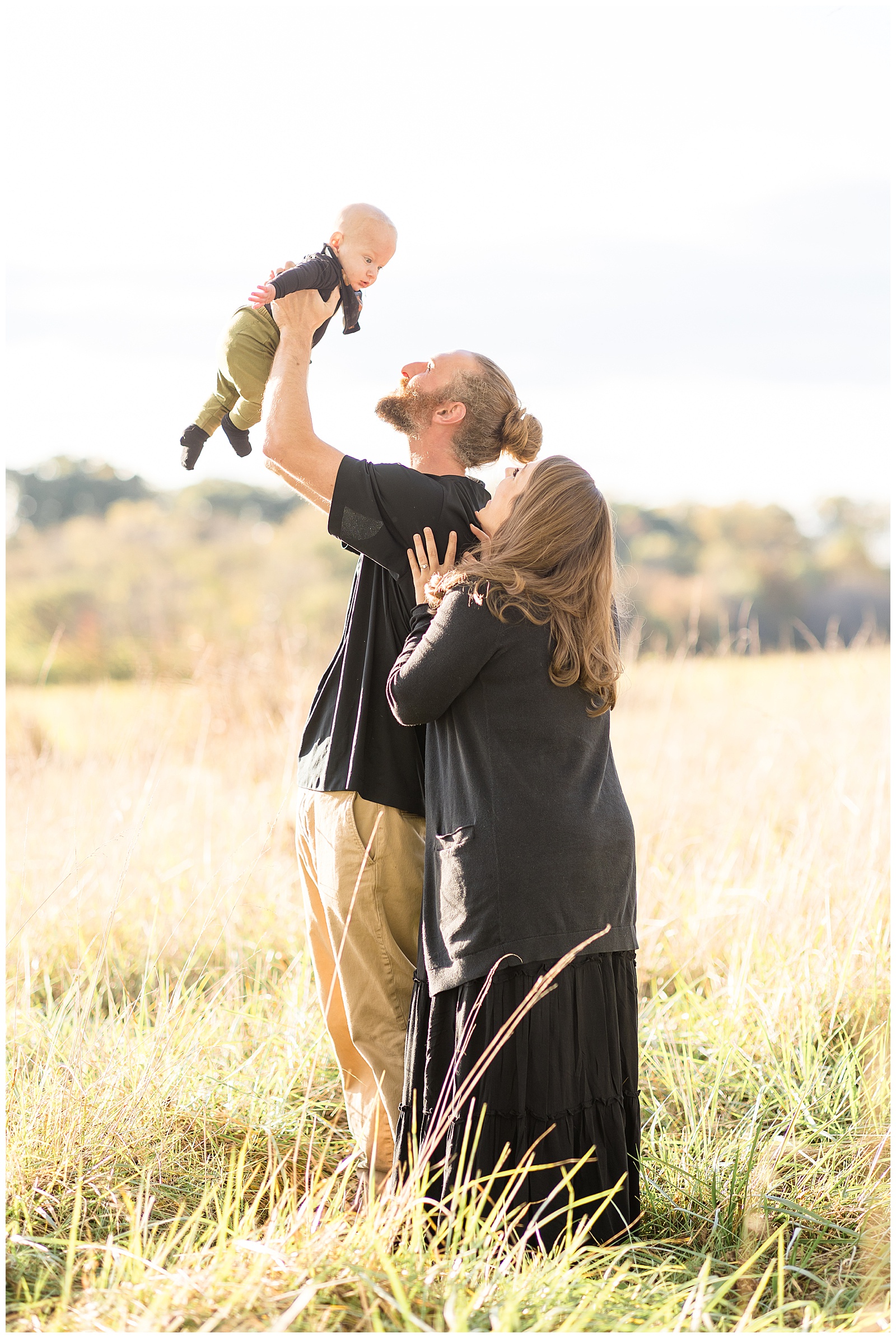 Dad holds baby boy up in the air as mom comes from behind his shoulder and they both look up at their son during their family photography session in Richmond, VA.