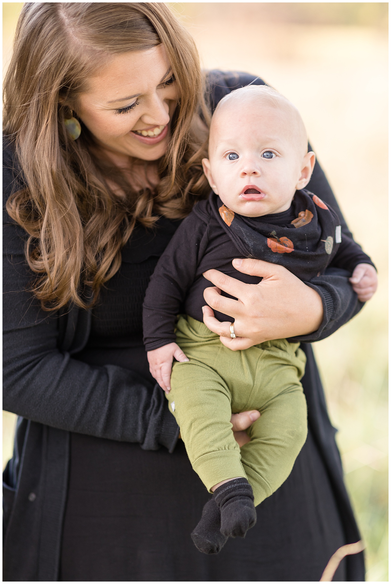 Mom, who is wearing a black dress and black cardigan, holds baby son who is wearing green pants and a black shirt with a pumpkin bib as he looks at camera with baby blue eyes.