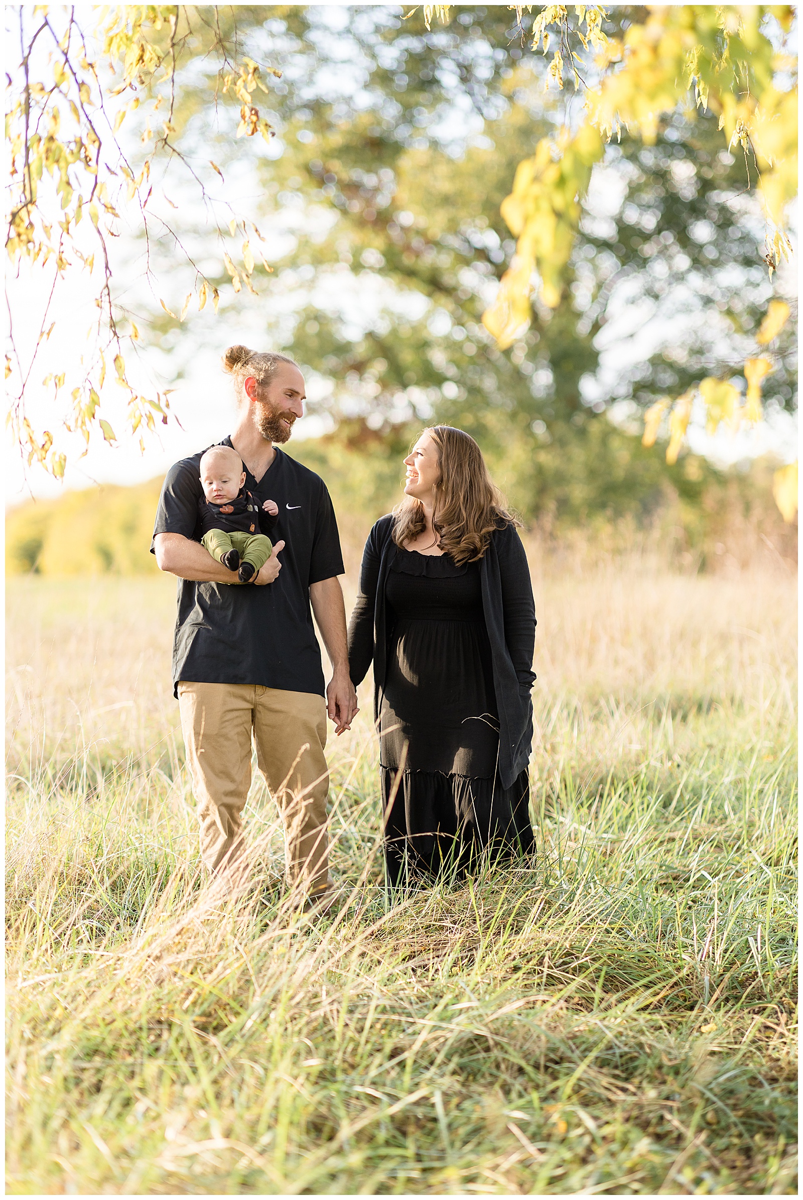 Husband and wife look at each other as they walk through a field in Richmond, VA with their baby boy who is being held by Dad.