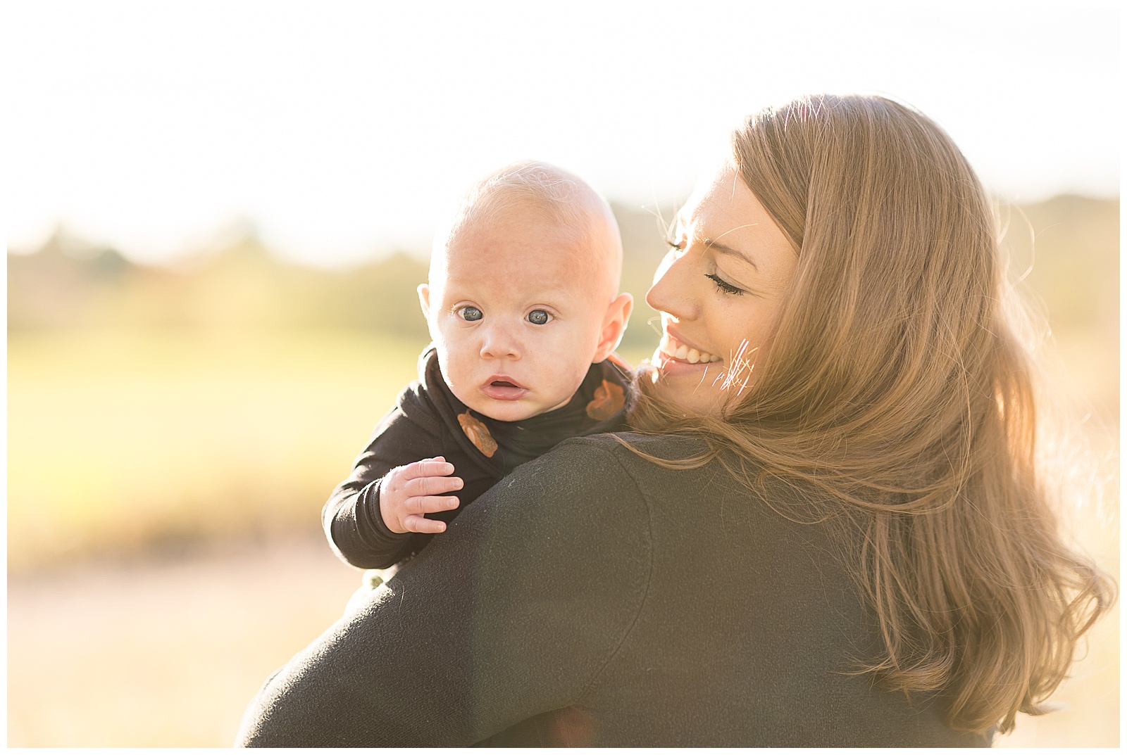 Mom holds baby boy over her shoulder who looks at the camera with his baby blue eyes and a sun flare coming through.