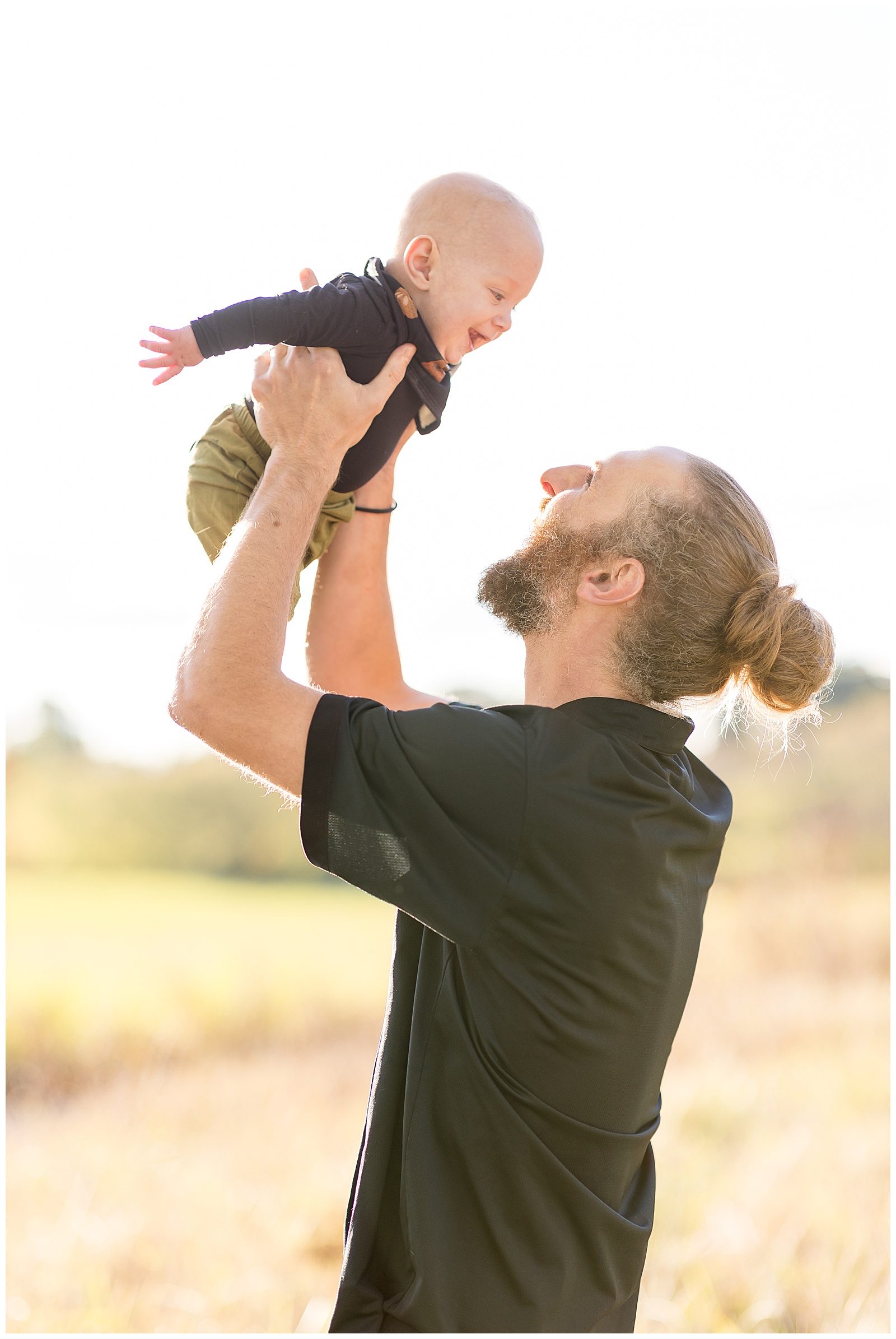 Dad, who has blonde hair and wears it in a bun, has on a black shirt and holds his baby boy up in the air as they smile at each other, both wearing black shirts.