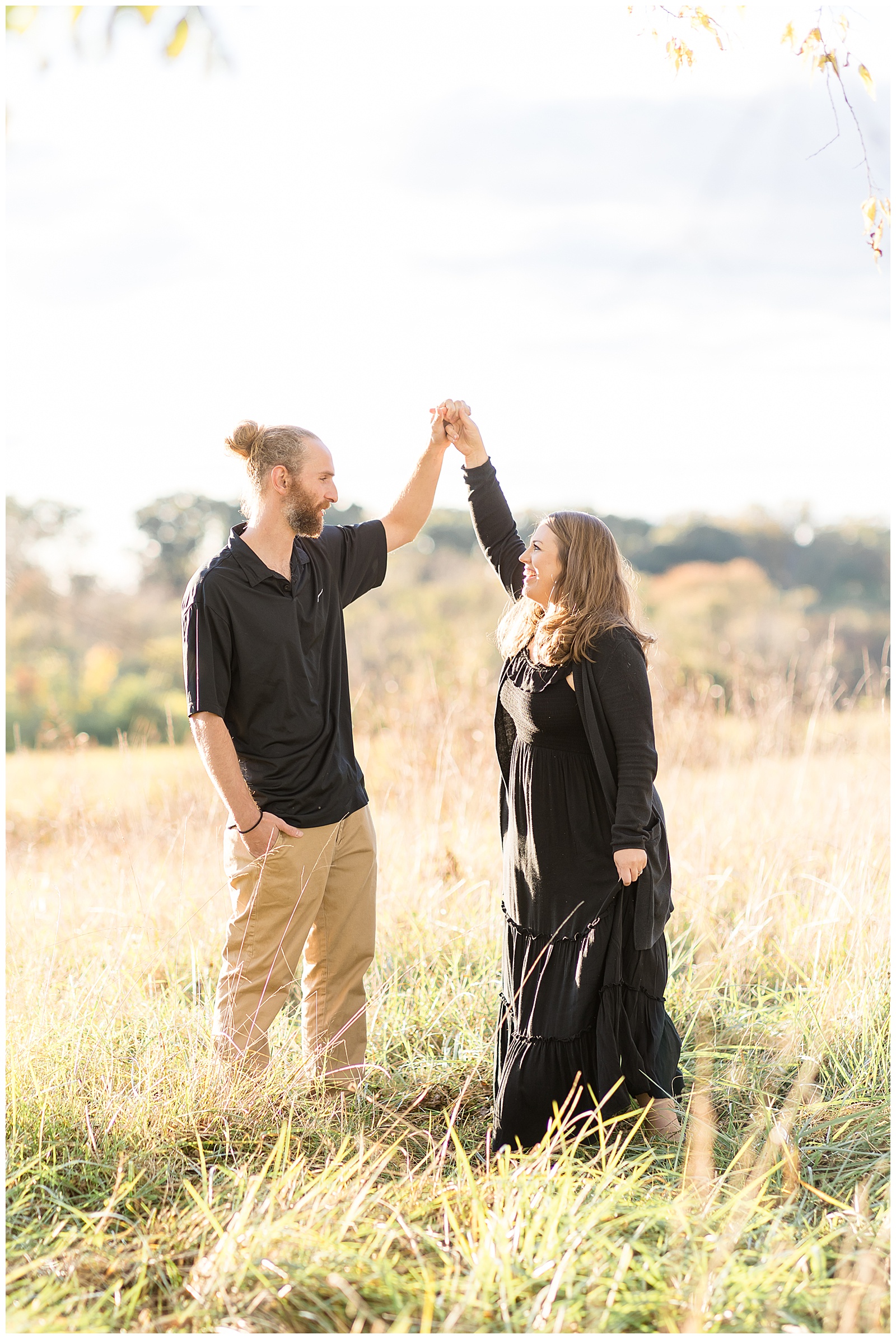 Couple dances in Richmond, VA in a field as the wife wears a black, long, dress and cardigan and her husband wears a black shirt and khaki pants as he twirls her in field.