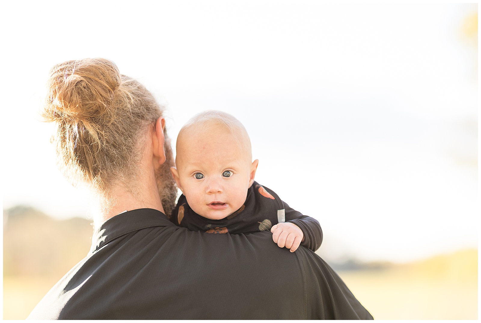 New Dad wears a blonde hair and bun with a black shirt and holds his baby boy over his shoulder as he looks at camera of Rebecca Rice with his baby blue eyes.