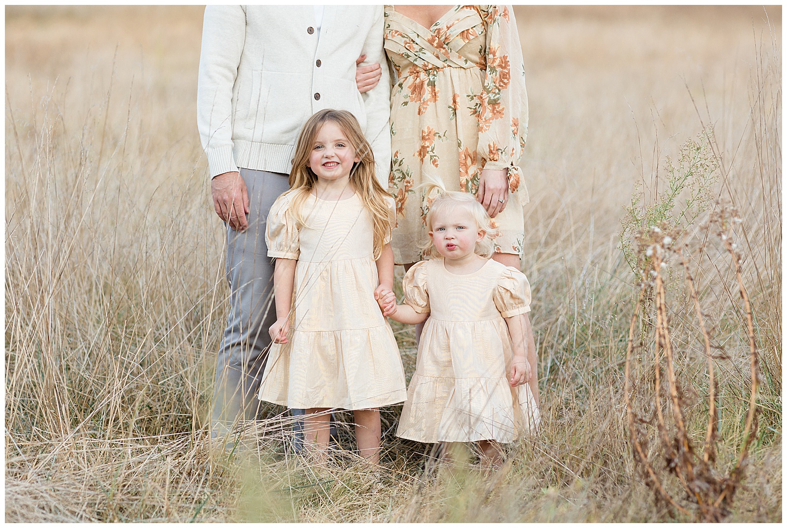Two young sisters wearing yellow dresses hold hands and smile at the camera as they stand in front of their parents during their family session in Nashville.