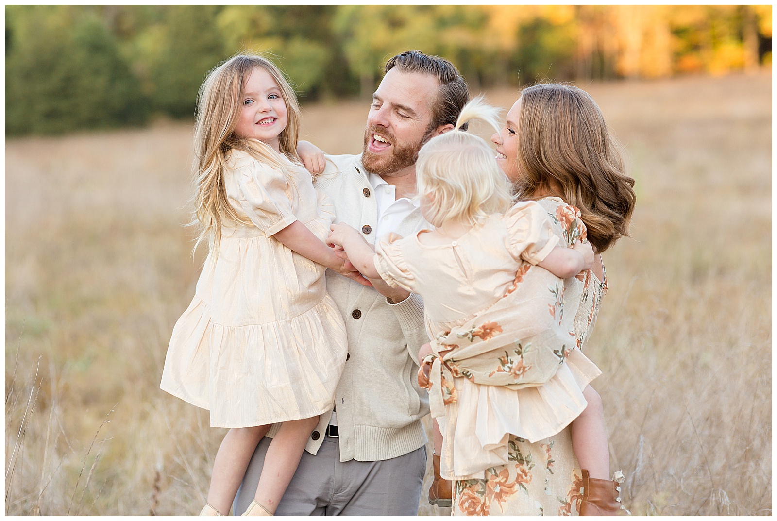 Mom and Dad hold their two young daughters as they laugh and tickle each other in a field in Nashville wearing coordinating yellow/cream colors.