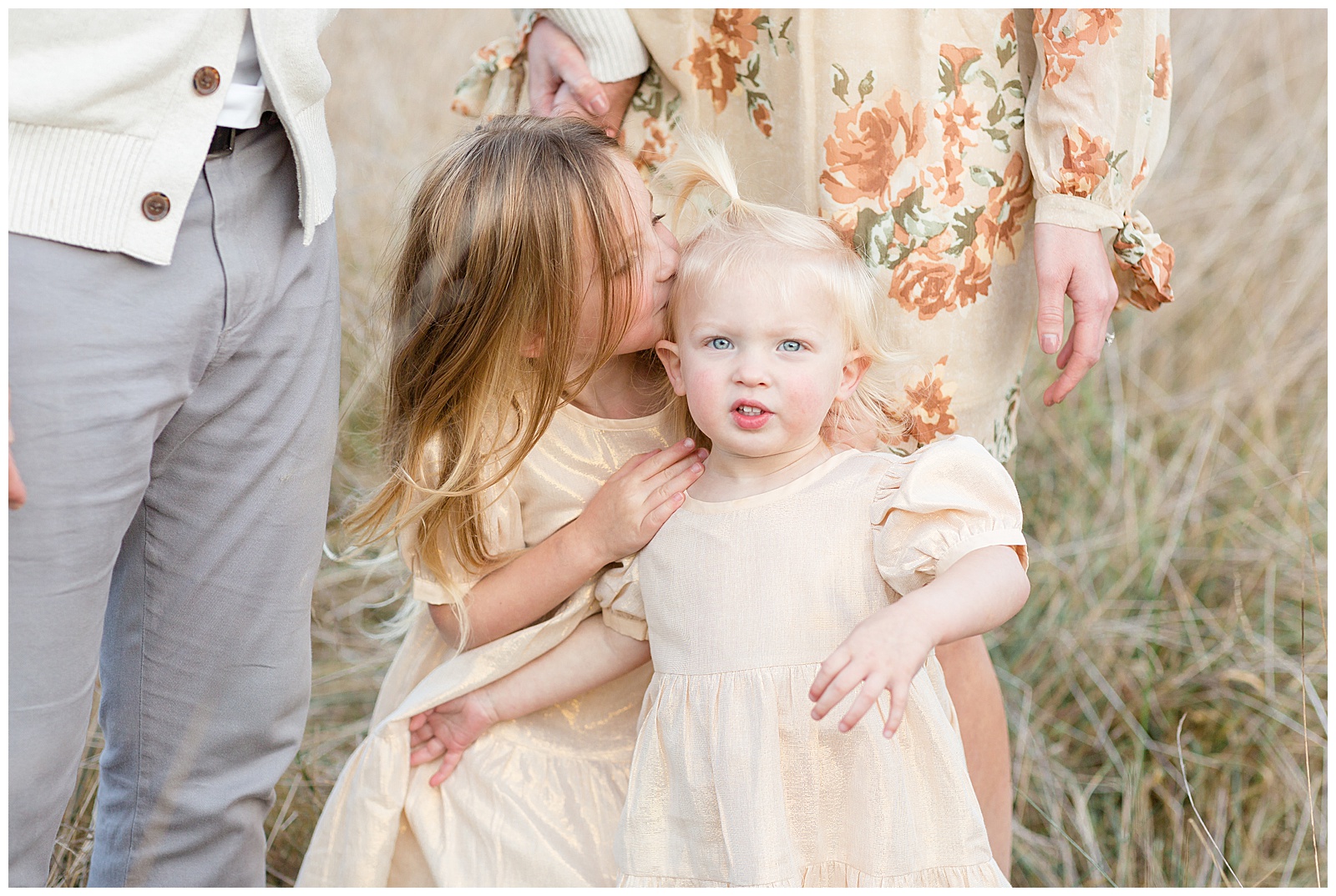 Two sisters stand in front of their parents which are not seen in the frame, wearing matching gold shimmery dresses and big sister gives a kiss on her little sisters head as the little one looks at the camera.