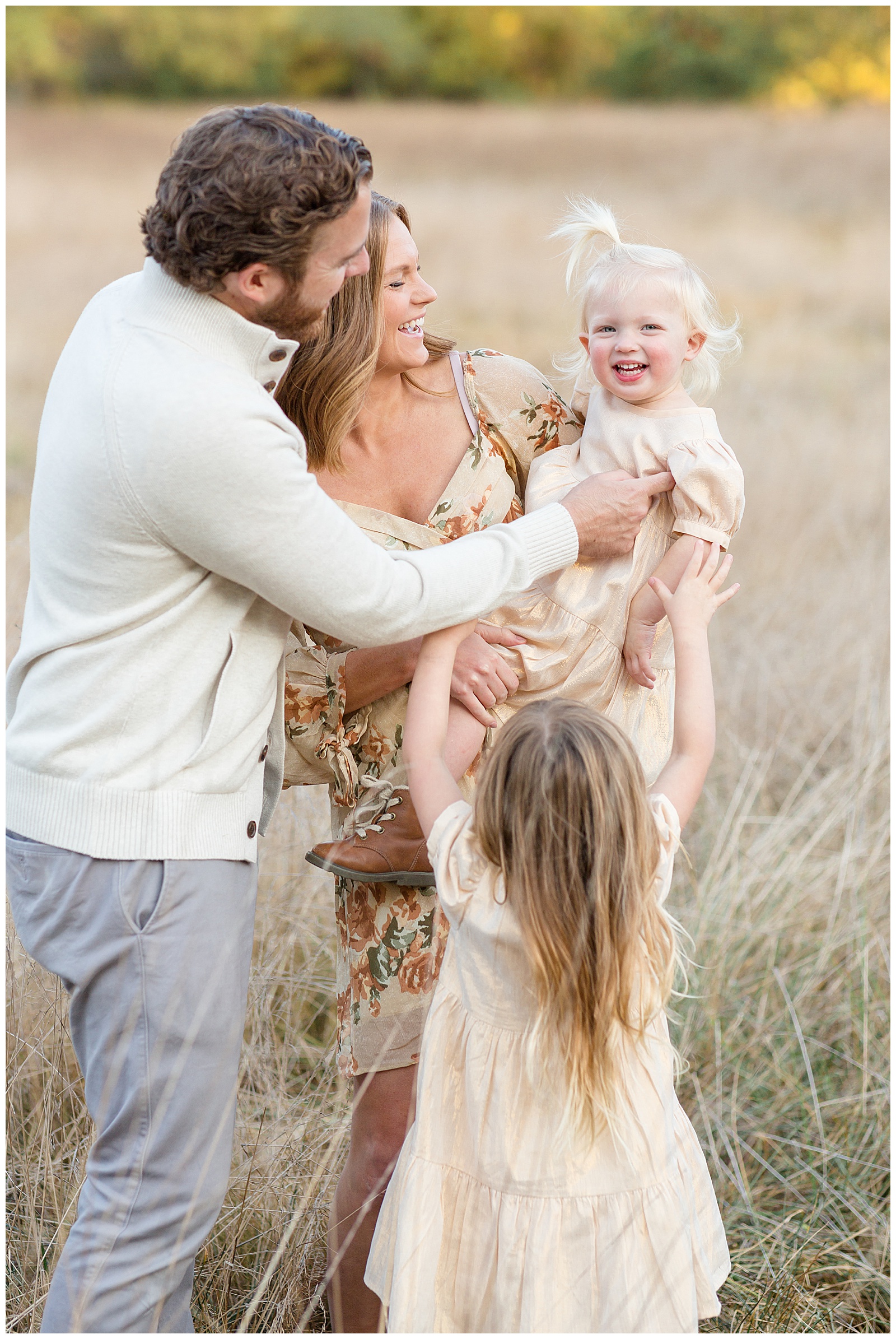 Family of 4 with two young daughters wearing coordinating color outfits of white and cream with mom wearing floral.  Mom holds youngest toddler daughter as big sister and dad all tickle her and she smiles big at the camera.  Click to see more on the blog today!