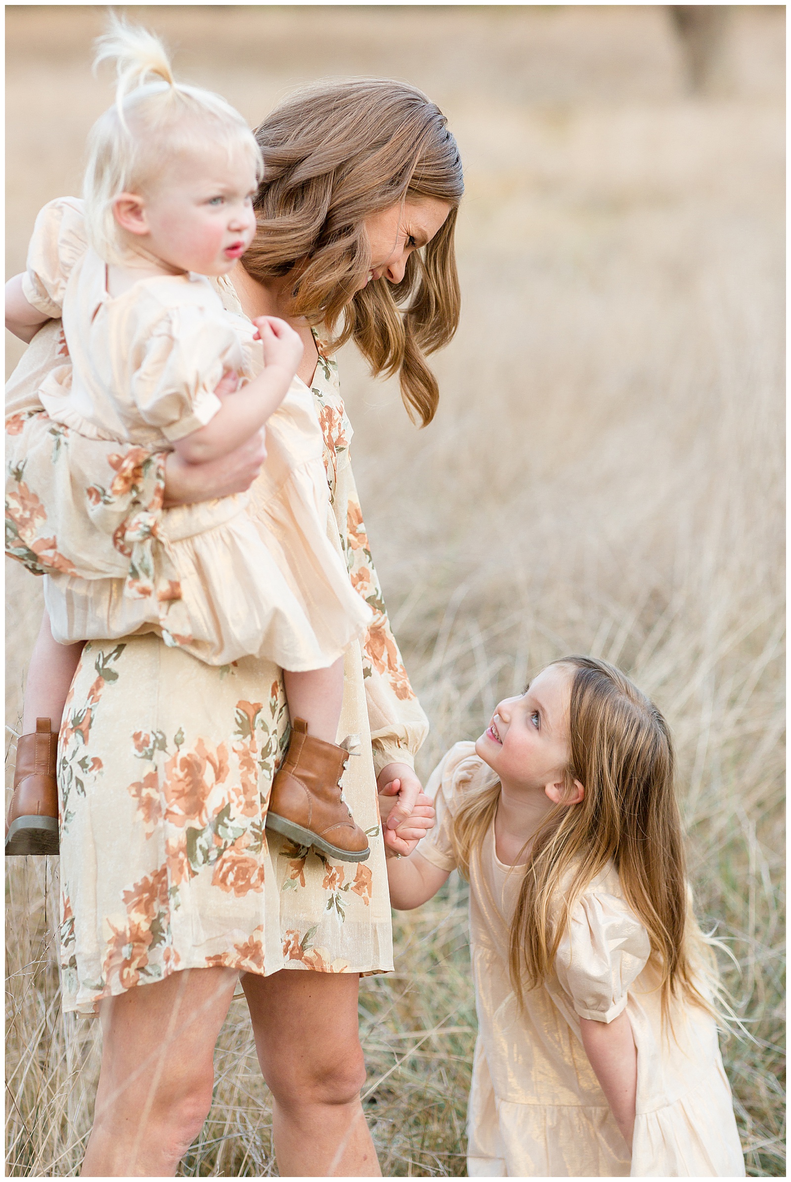 Mom is wearing a cream colored, bohemian floral v-neck dress as she holds her toddler daughter and looks down at her oldest daughter and holds her hand as they look and smile at each other.