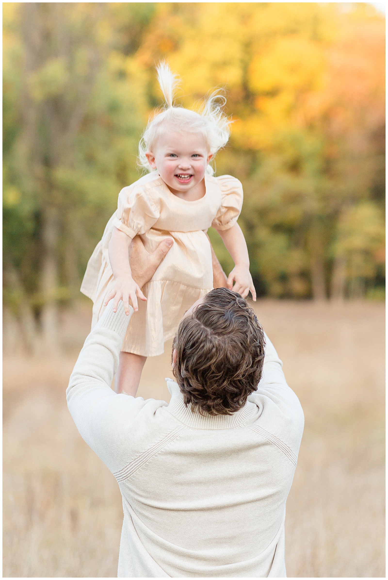 Dad, wearing a white sweater has his back turned to the camera as he lifts his toddler daughter up in the air as she smiles at the camera with her gold, shimmery dress.