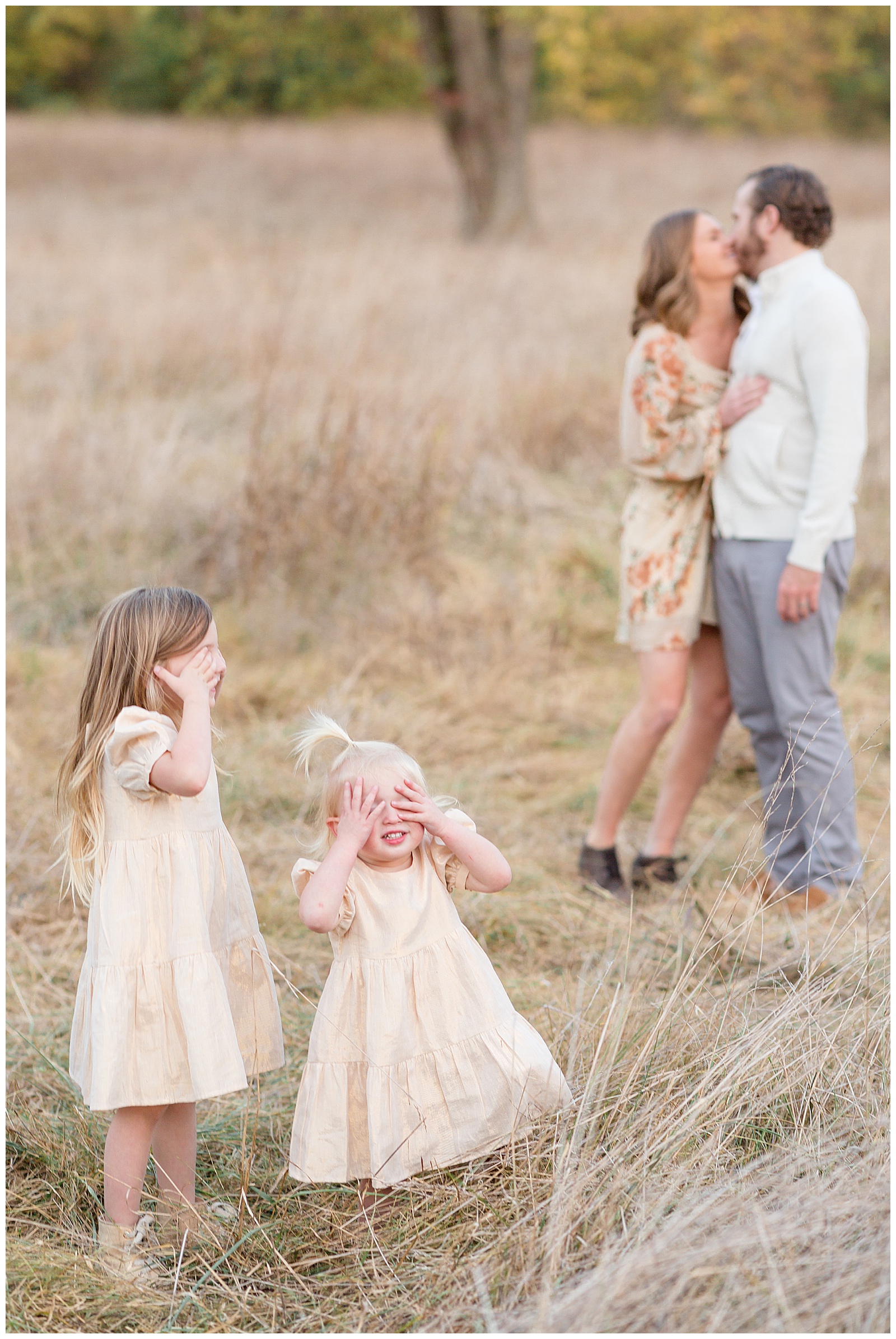 Mom and Dad share a kiss in the Nashville field in the background as their two toddler daughters stand in the forefront with matching gold dresses covering their eyes.