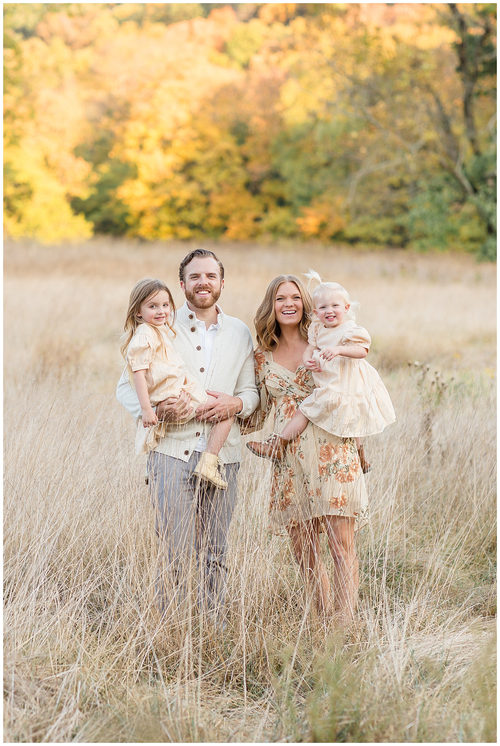 Family of 4 with two toddler daughters, are being held by Mom and Dad in a field in Nashville during their family session.  They wear neutral colors that blend in beautifully with the tall grass field as the sun highlights the trees behind them.