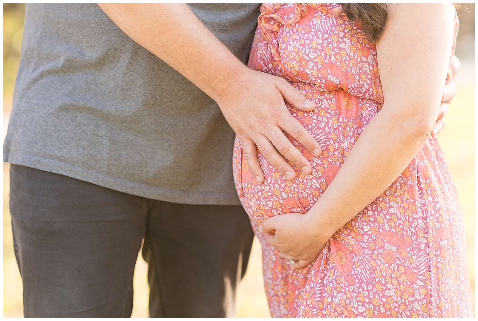 Detail photo of husband holding his pregnant wife's belly and she holds underneath her belly and wears a pink, orange, and peach designed dress.