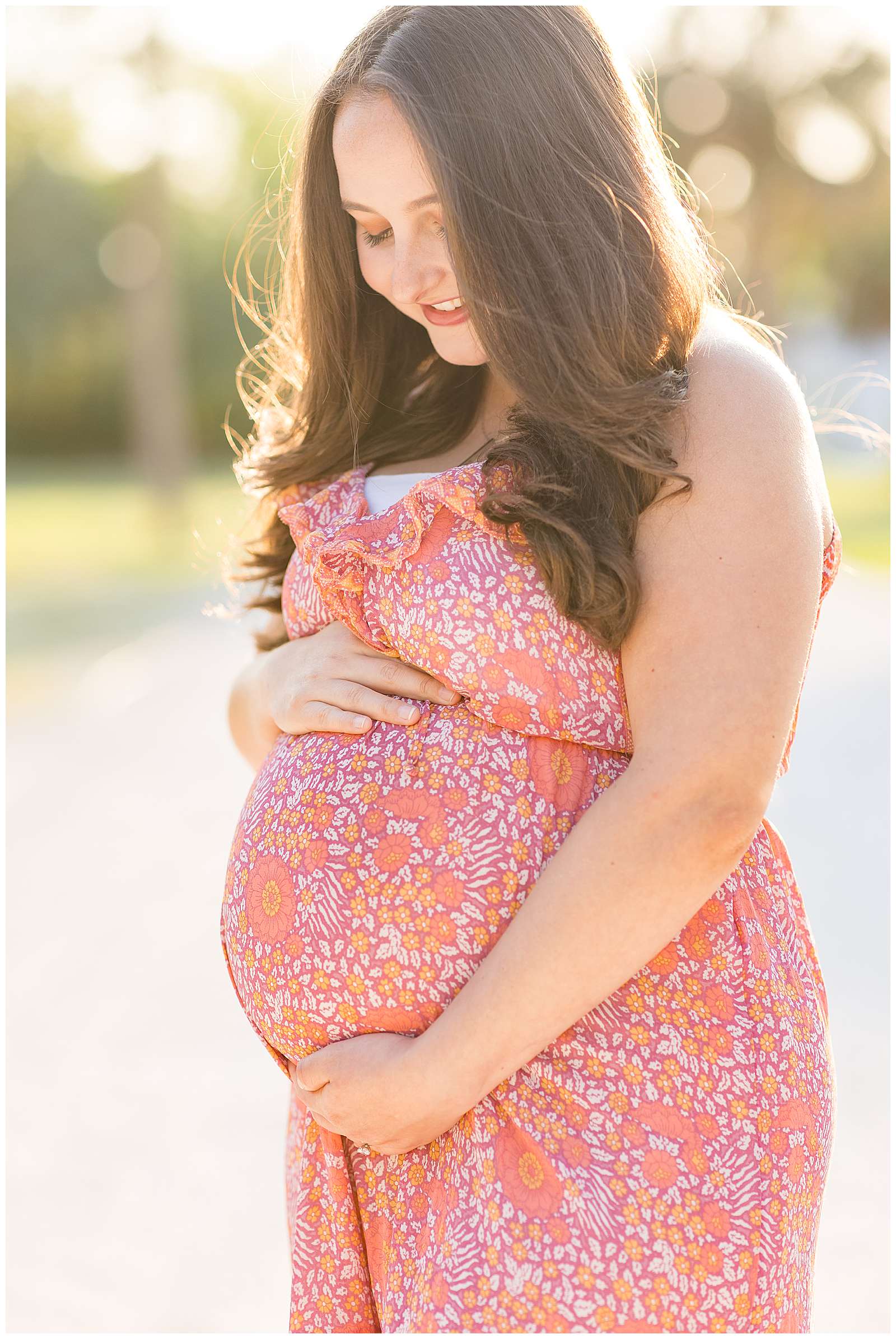 Pregnant momma holds her belly and looks down at her baby on the way!  She wears a pink, orange, and peach tone designed dress.