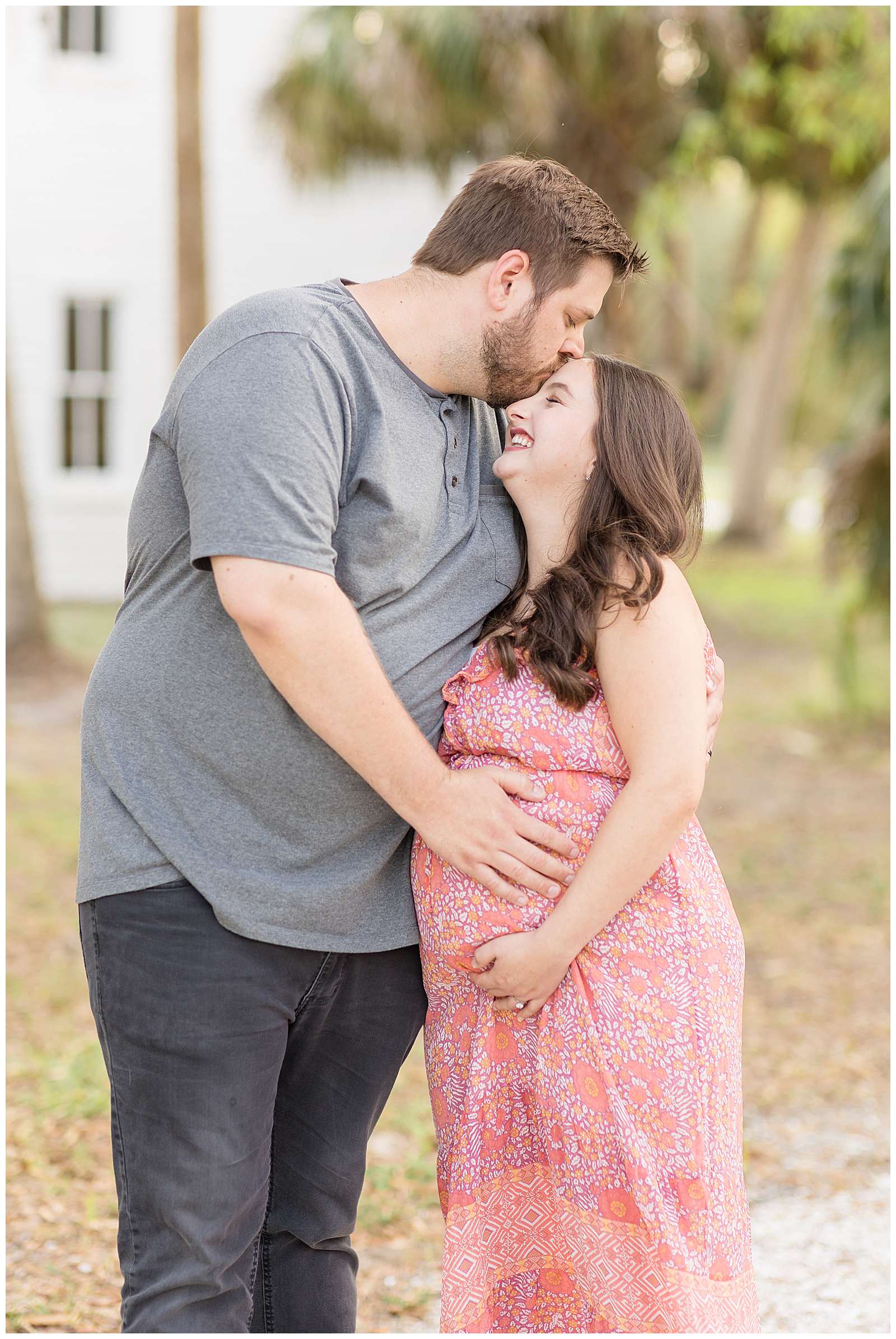 The sweetest couple with the biggest height difference share a laugh during their Florida maternity session with Rebecca Rice Photography.  Husband leans down to kiss his wife on her forehead and she looks up with her eyes closed laughing.