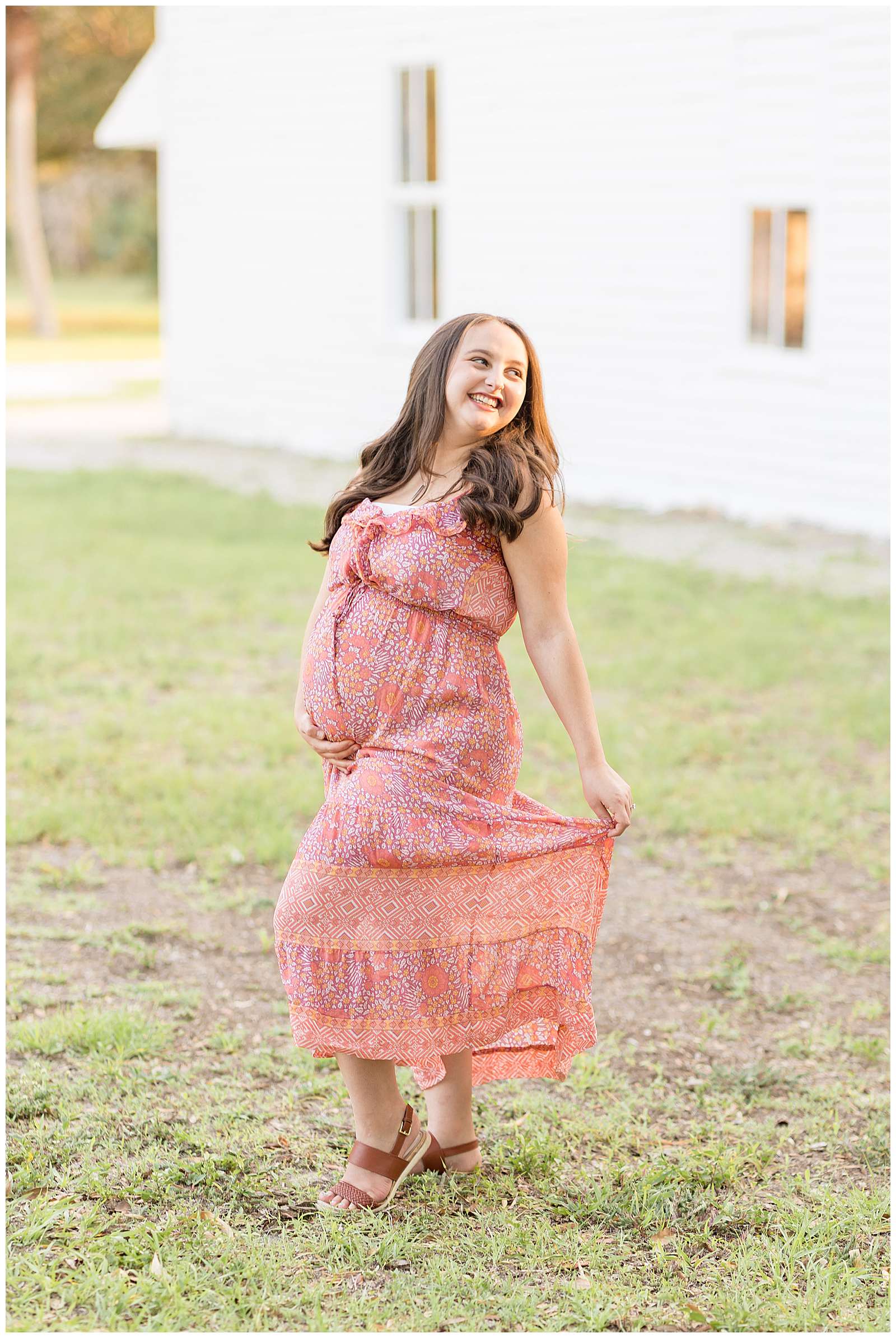 Pregnant momma stands in the grass in front of a white building and looks off over her shoulder as she smiles in the distance.  She wears a pink, orange, peach colored, designed dress.