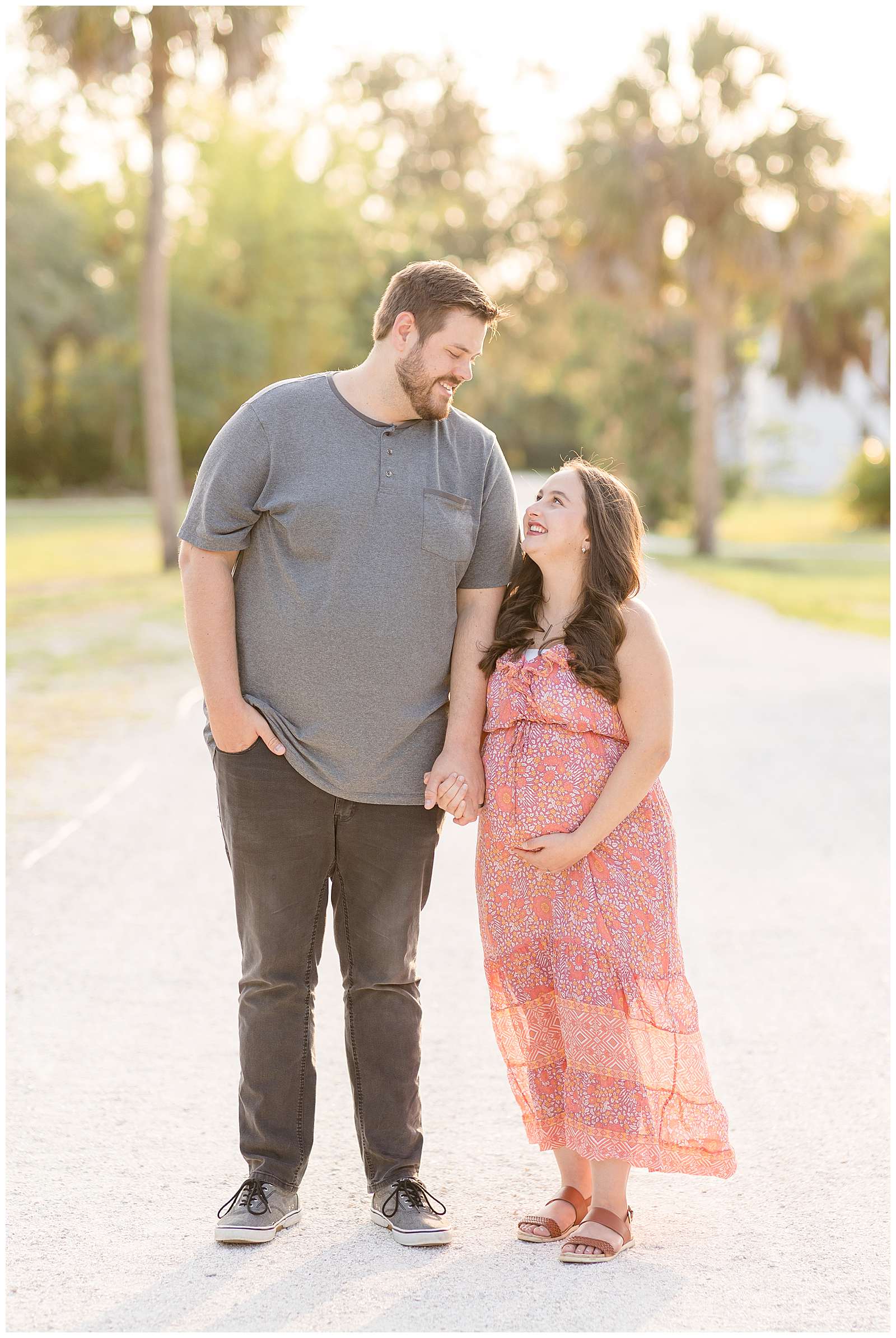 Rebecca Rice Photography captures a BIG height difference maternity session for her Behind the Lens membership.  The husband wears a grey t-shirt and dark grey jeans.  He holds his wife's hand as he looks down at his wife who smiles up at him holding her belly.
