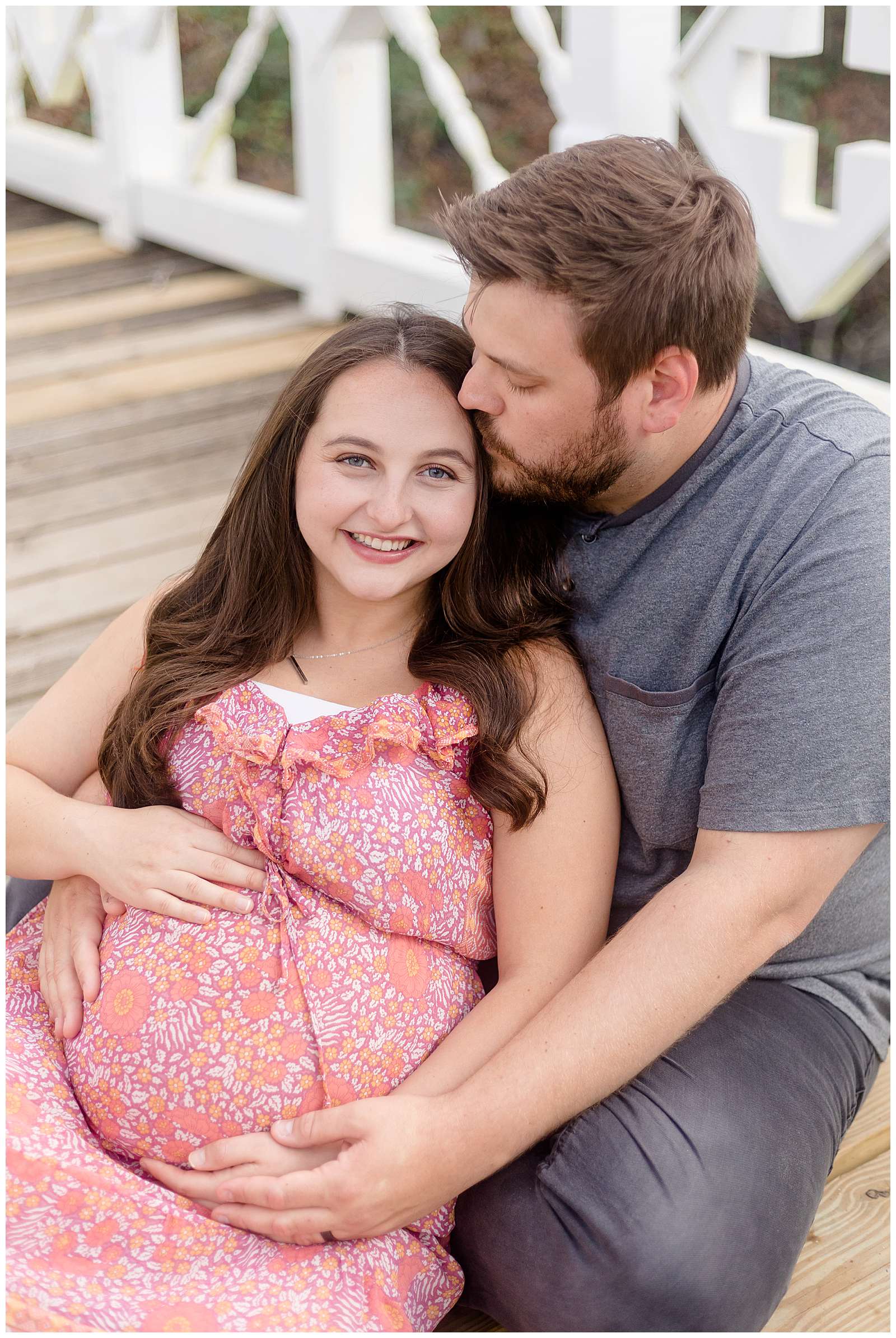 Pregnant momma who is wearing a pink, orange, and peach colored designed dress leans back into her husbands lap as he leans forward and helps hold her pregnant belly.  They sit on a bridge in Florida during their maternity session.