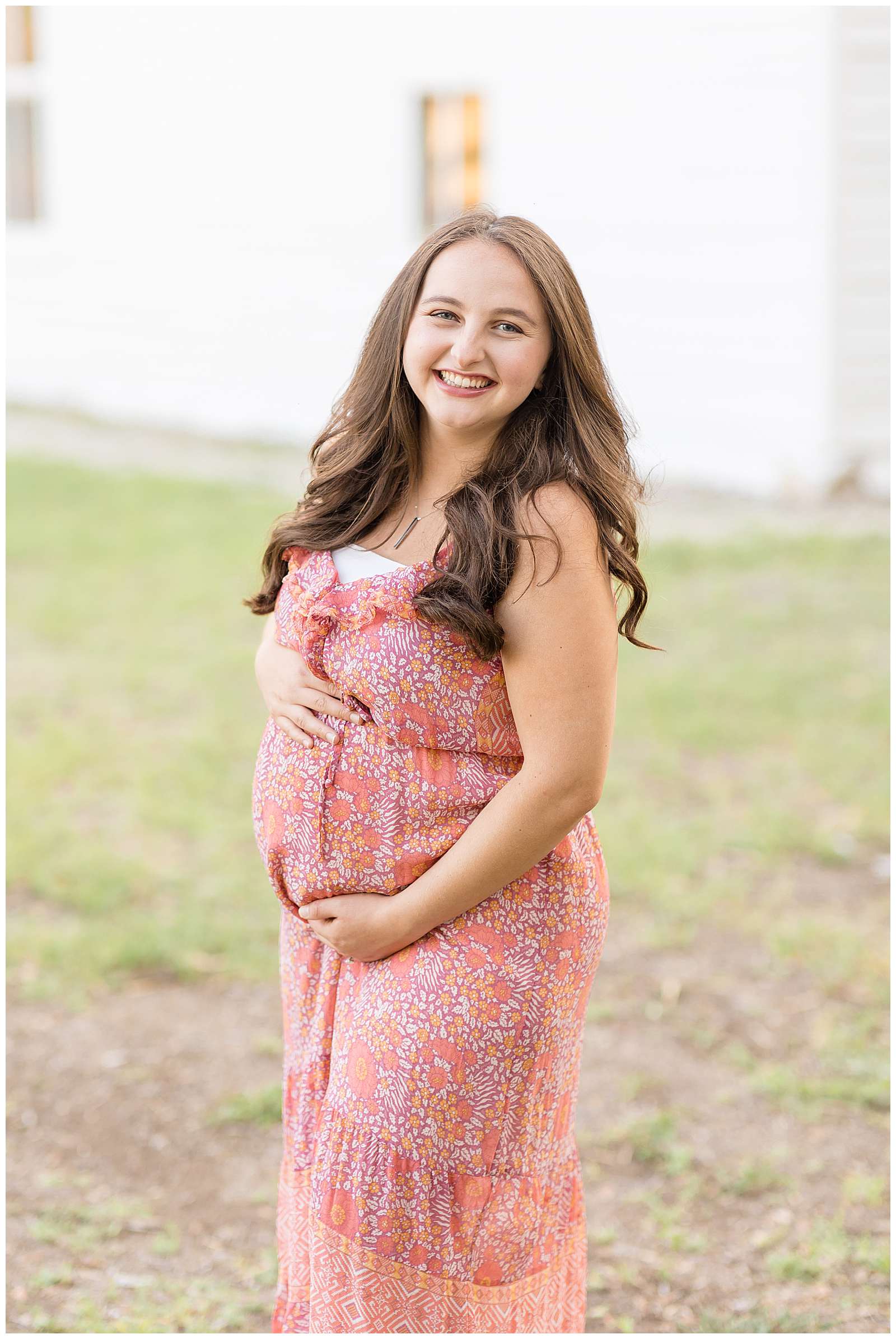Beautiful pregnant momma smiles at the camera of Rebecca Rice Photography wearing a bright, summer dress and holds her belly as she leans slightly forward.