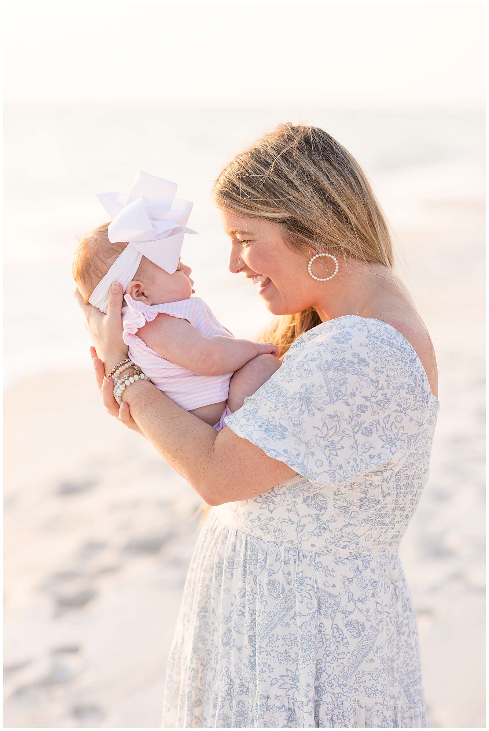 New mom who is wearing a white and blue floral dress with hoop earrings and bracelets, holds her baby girl in her arms sitting on her chest wearing a pink and white short sleeve and shorts outfit and a big, white headband bow.  Click to see more on the blog today! -rebeccaricephoto.com