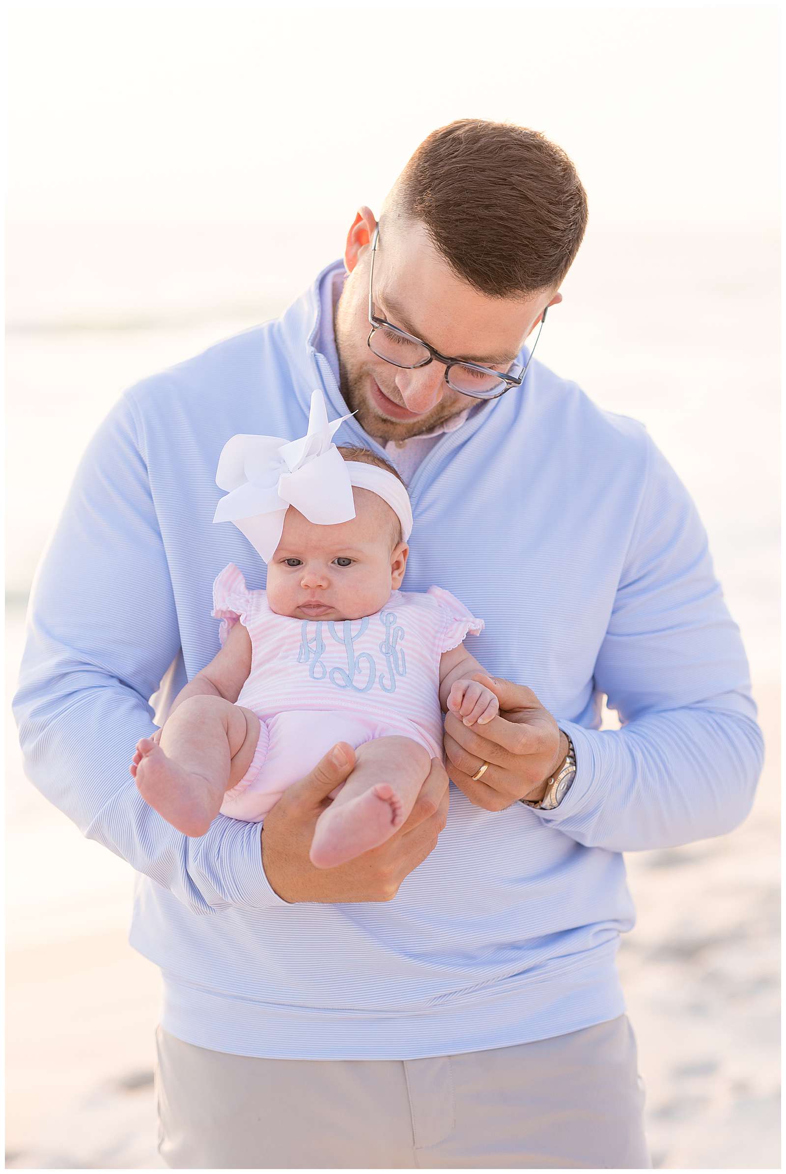 New Dad who wears a light blue half zip long sleeve shirt and khakis, holds his baby girl with one hand facing out and the other holding her little arm.  The baby wears a pink and white stripped, monogrammed onesie and a big white headband bow at Rosemary Beach.  Click to see more on the blog today! -rebeccaricephoto.com
